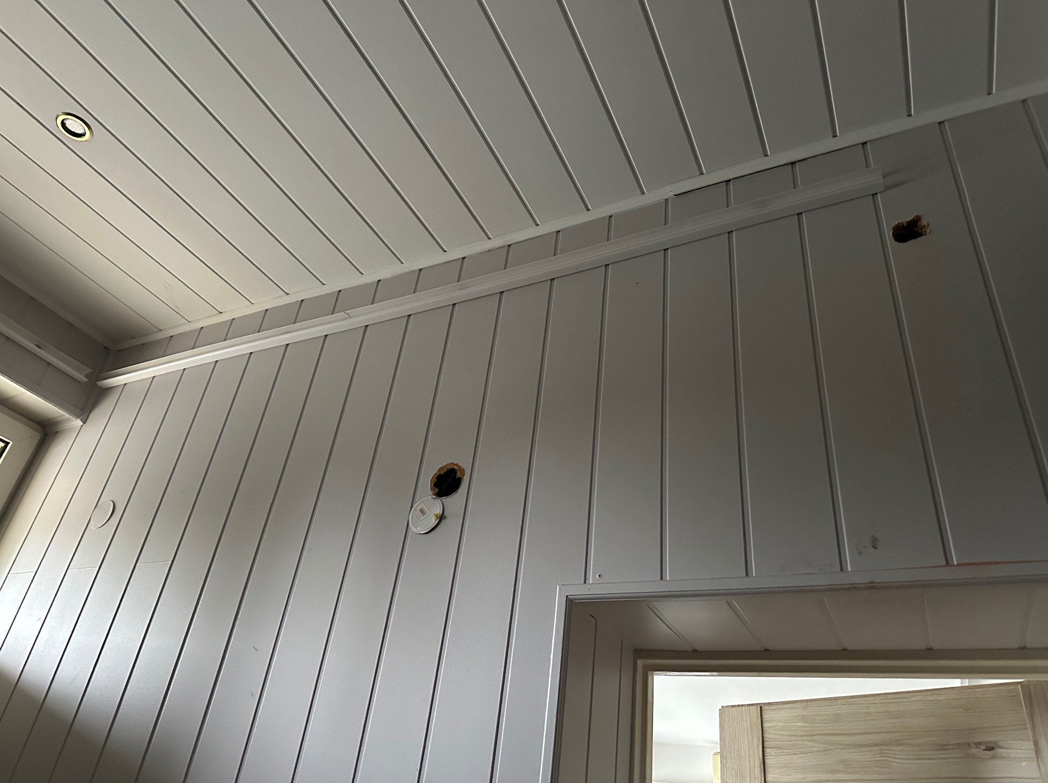 Szare panele ścienne i sufitowe z widocznymi otworami na oświetlenie i gniazdka. Częściowo zamontowana listwa dekoracyjna. Widoczny fragment drzwi.