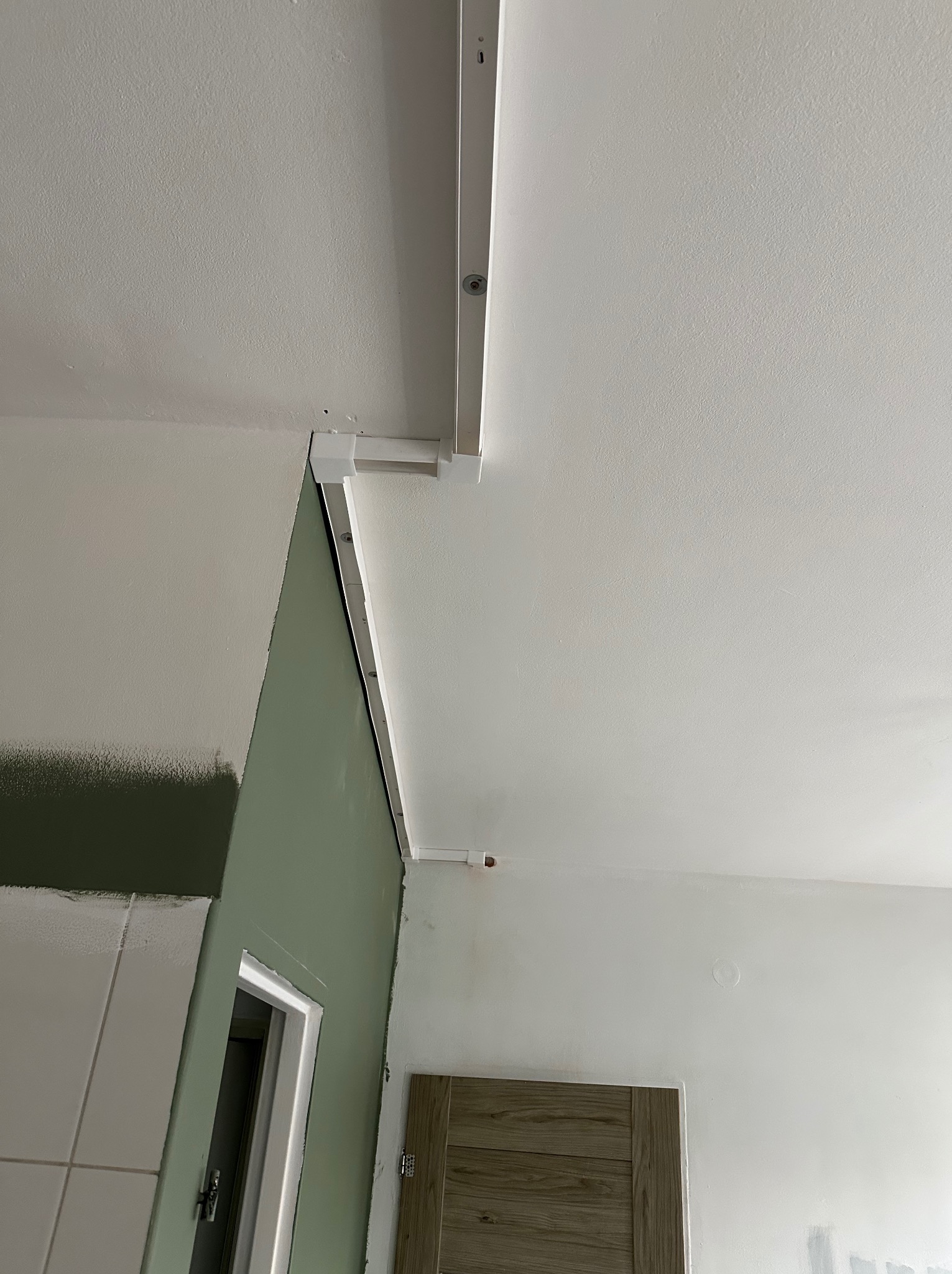 Montaż szyny oświetleniowej na suficie, widoczne przewody elektryczne i fragment ściany z świeżo nałożoną farbą w odcieniach zieleni. Prace wykończeniowe w trakcie.