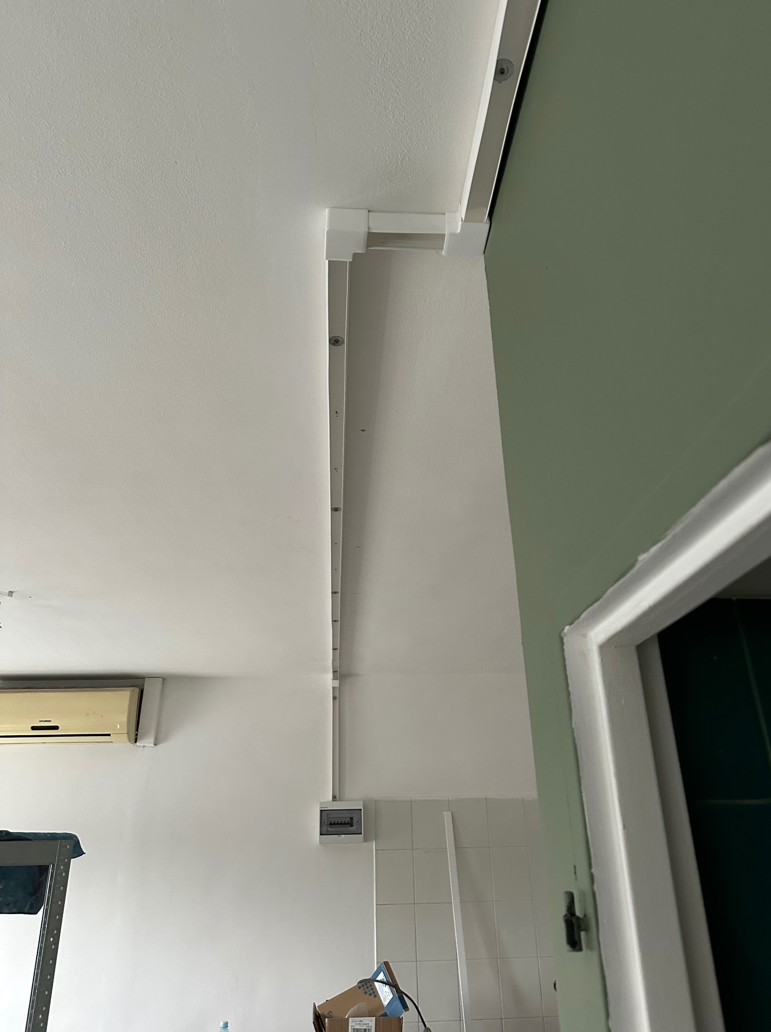 Biała listwa sufitowa z zamontowanymi punktami świetlnymi, widoczna na tle białego sufitu i fragmentu zielonej ściany. Widoczna skrzynka elektryczna i klimatyzator.