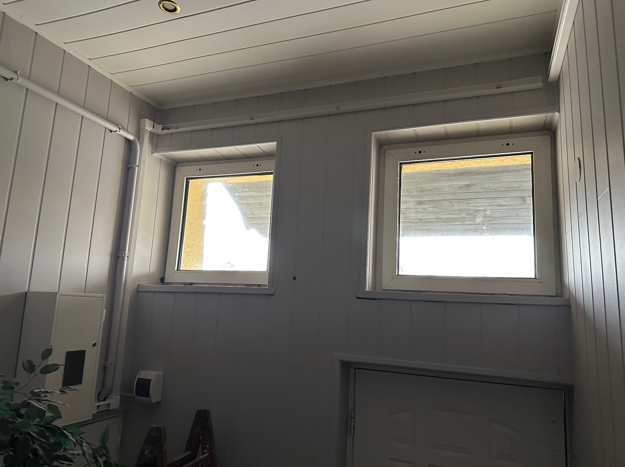Wnętrze wykończone panelami ściennymi w odcieniach bieli, z dwoma oknami z roletami i widoczną skrzynką elektryczną. U dołu fragment drabiny i drzwi.
