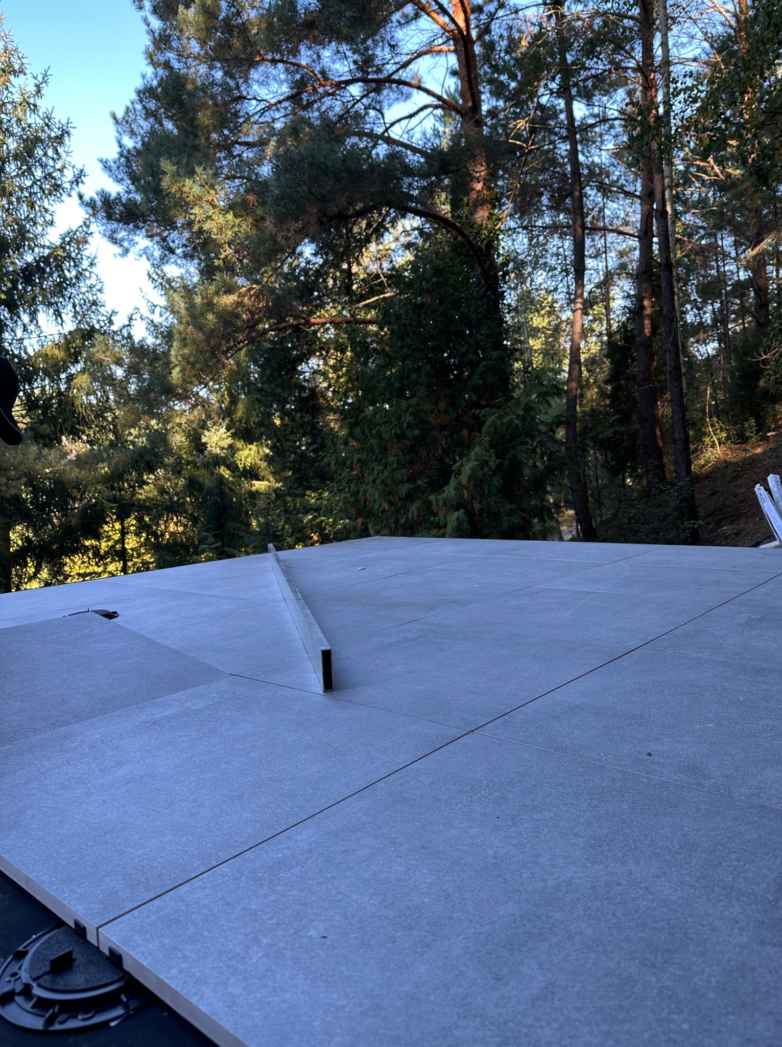 Układanie szarych płyt tarasowych na regulowanych wspornikach, widoczne drzewa w tle. Prace wykończeniowe na zewnątrz, poziomowanie powierzchni.