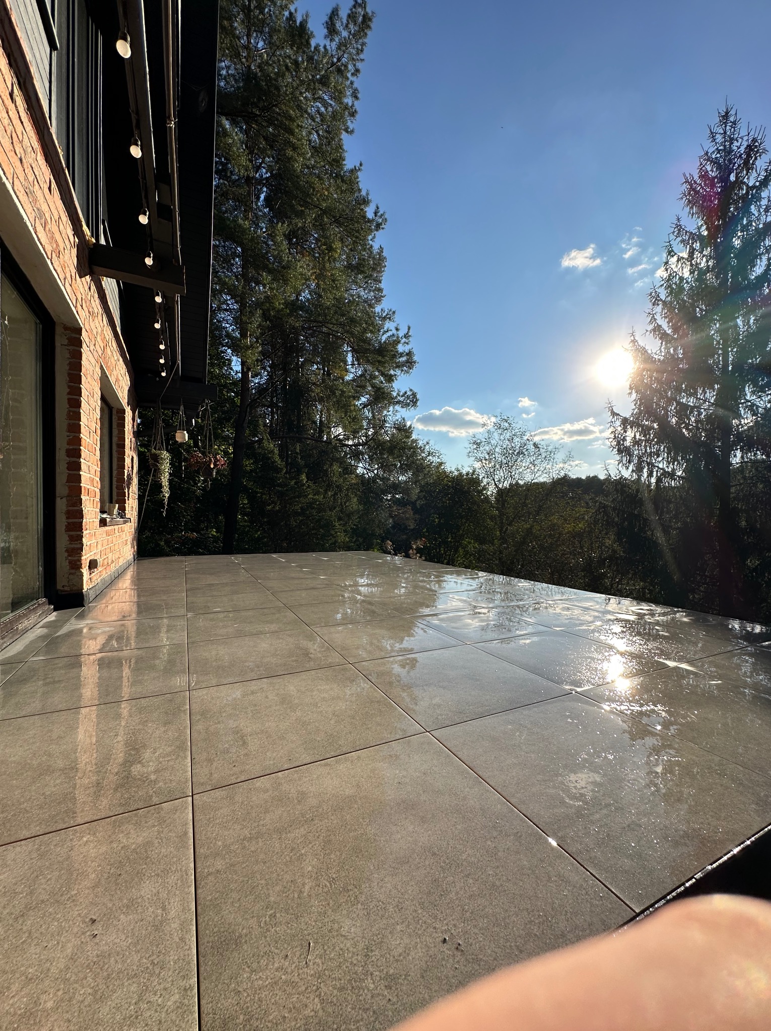 Wyłożony jasnymi płytkami taras z widokiem na zieleń, oświetlony słońcem. Widoczny fragment ceglanej ściany budynku z oknami i wiszącymi roślinami.