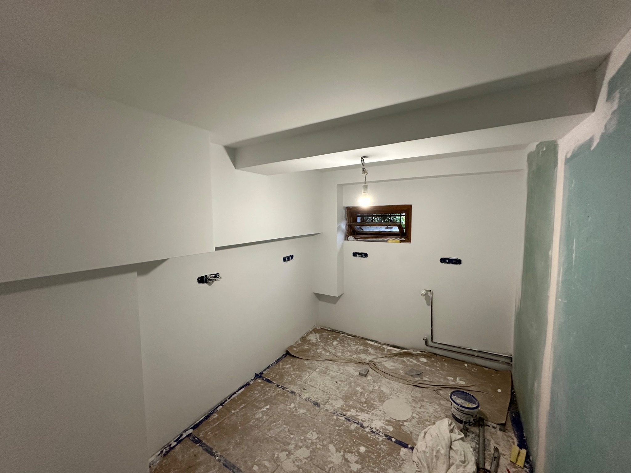 Wnętrze po malowaniu na biało, widoczne puszki elektryczne, okno z drewnianą ramą i zabezpieczona podłoga. Ściana po prawej częściowo pomalowana na zielono.