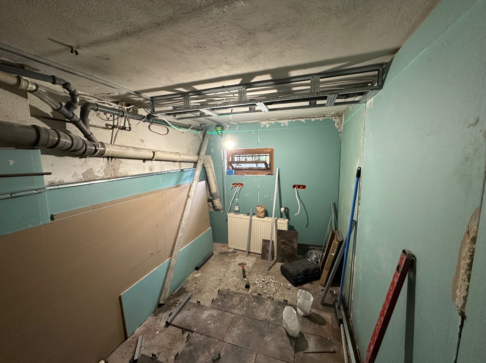 Wnętrze w trakcie remontu: widoczne rury, instalacja pod sufitem, płyty gipsowe i narzędzia. Ściany w kolorze miętowym, podłoga częściowo wyłożona płytkami.