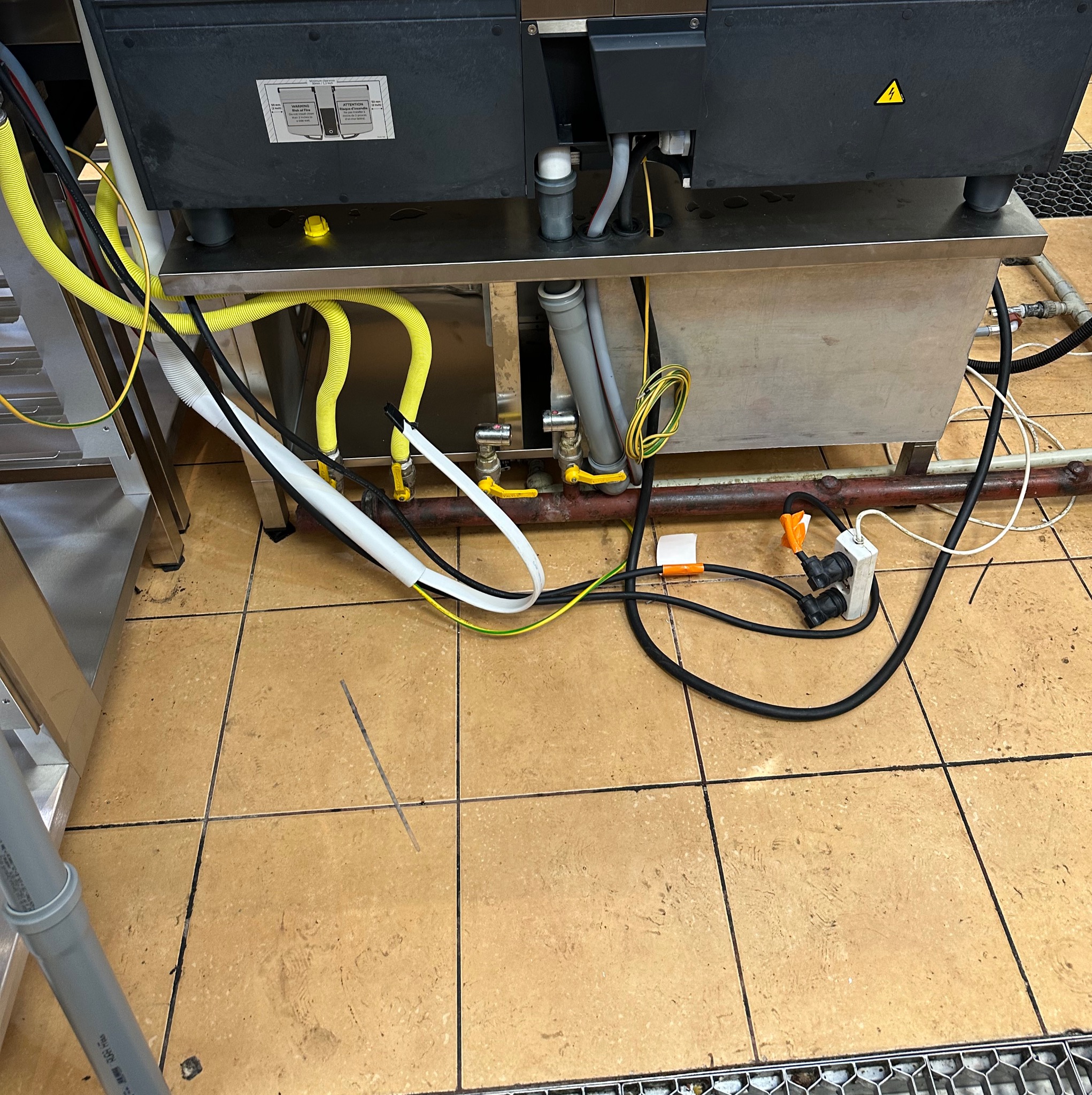 Szczegółowe ujęcie instalacji pod urządzeniem: żółte i białe przewody, szare rury PCV, czarne kable zasilające i gniazdka, widoczne na brązowej podłodze z płytek.