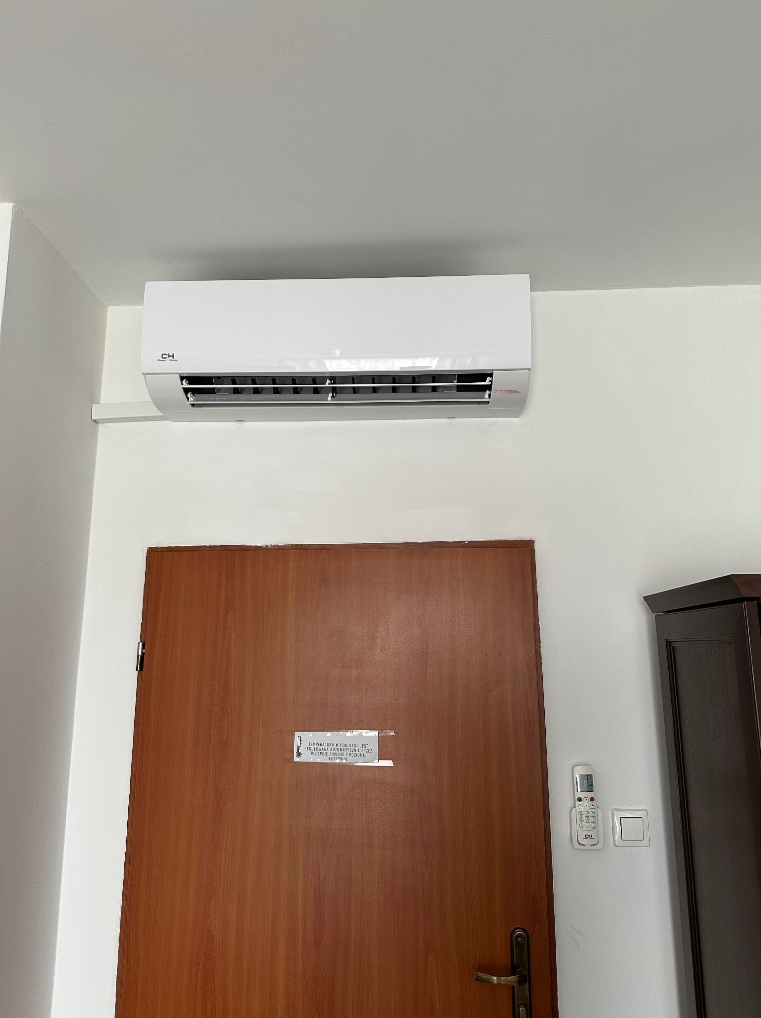 Biały klimatyzator ścienny marki CH nad brązowymi drzwiami z naklejką informacyjną i pilotem na białej ścianie. Widoczny fragment ciemnego mebla.