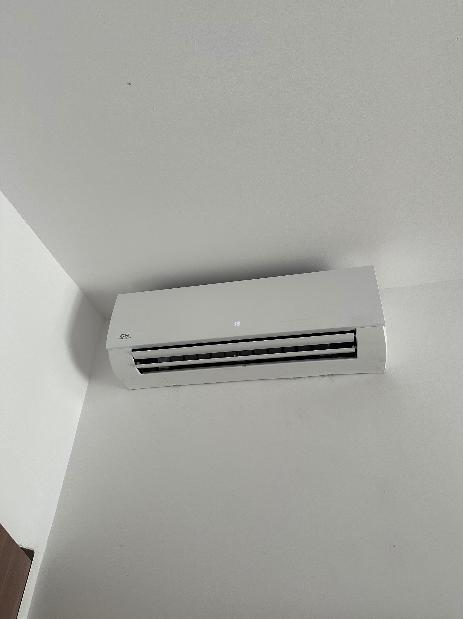 Biały klimatyzator ścienny marki CH, zamontowany w rogu pomieszczenia z białymi ścianami. Widoczny wyświetlacz z temperaturą 19 stopni.