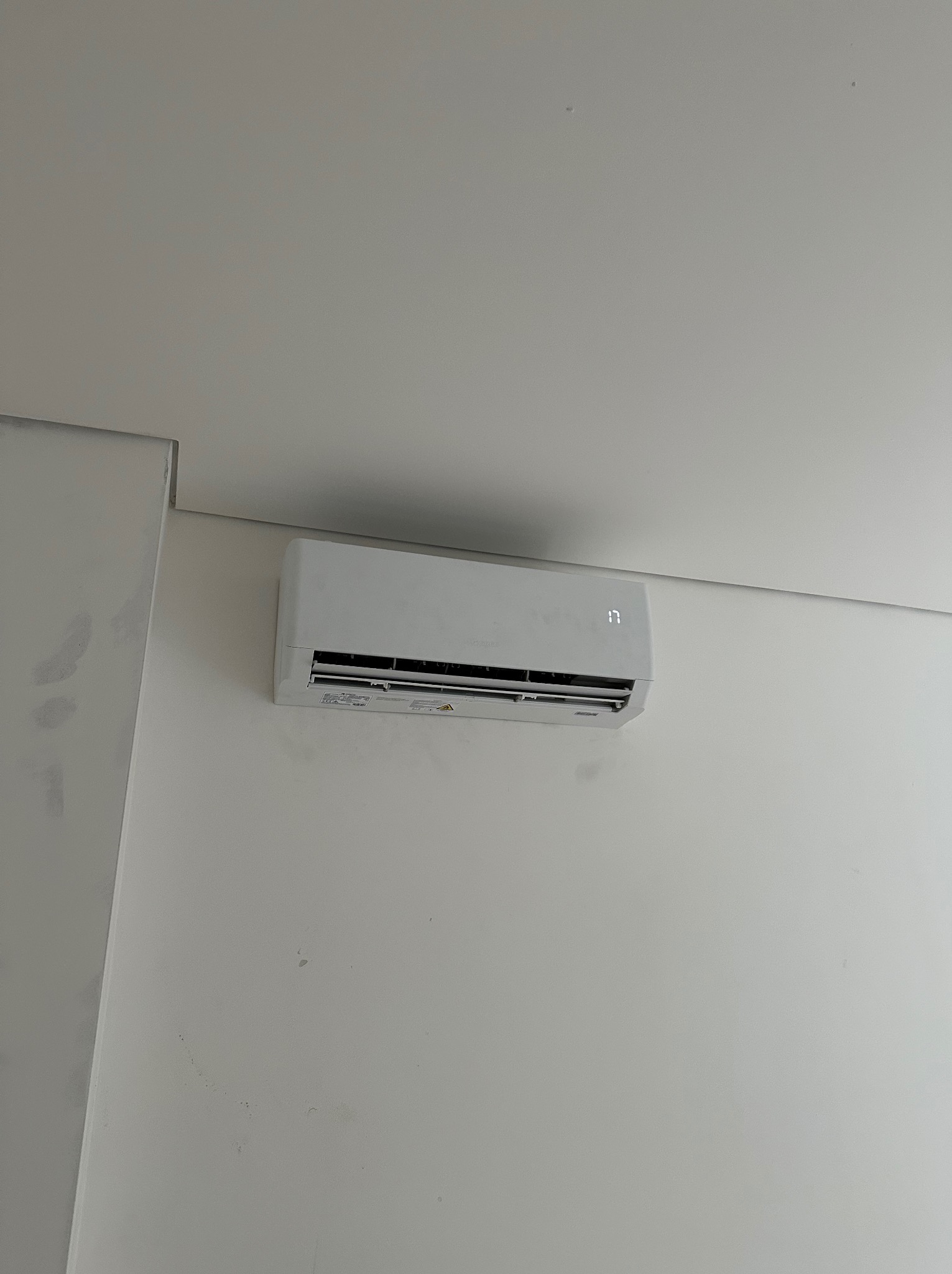 Biały klimatyzator ścienny z wyświetlaczem temperatury, zamontowany w rogu pomieszczenia o białych ścianach i suficie, widok z dołu.