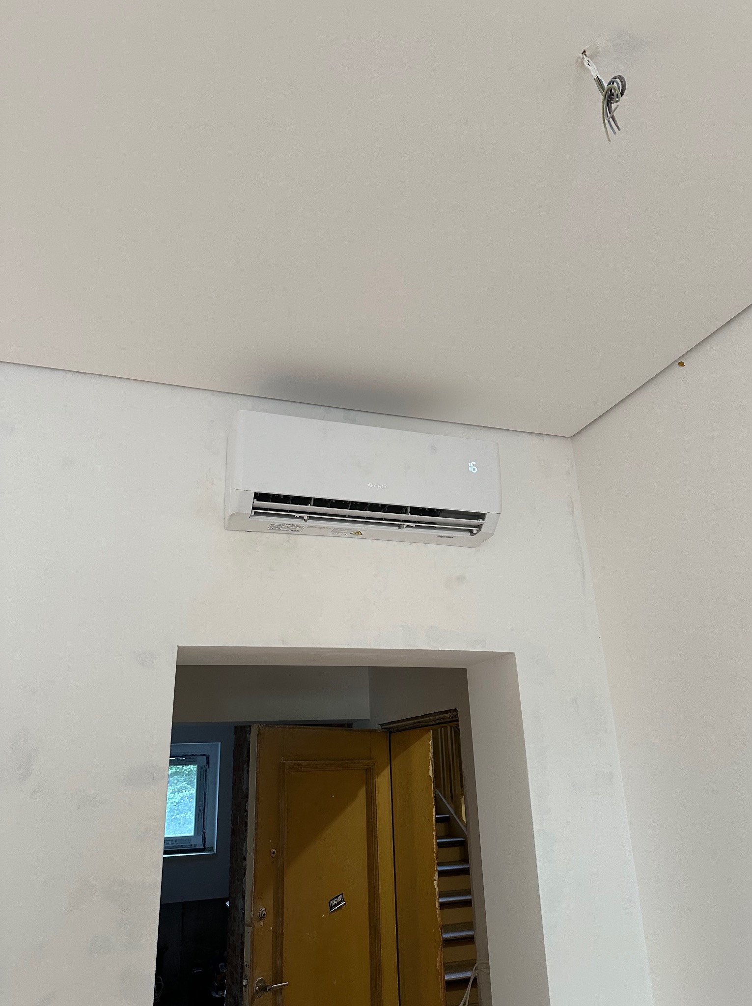 Biały klimatyzator na ścianie nad wejściem do pokoju. Widoczne niedokończone wykończenia ścian i wystające przewody elektryczne. Ujęcie z dołu.
