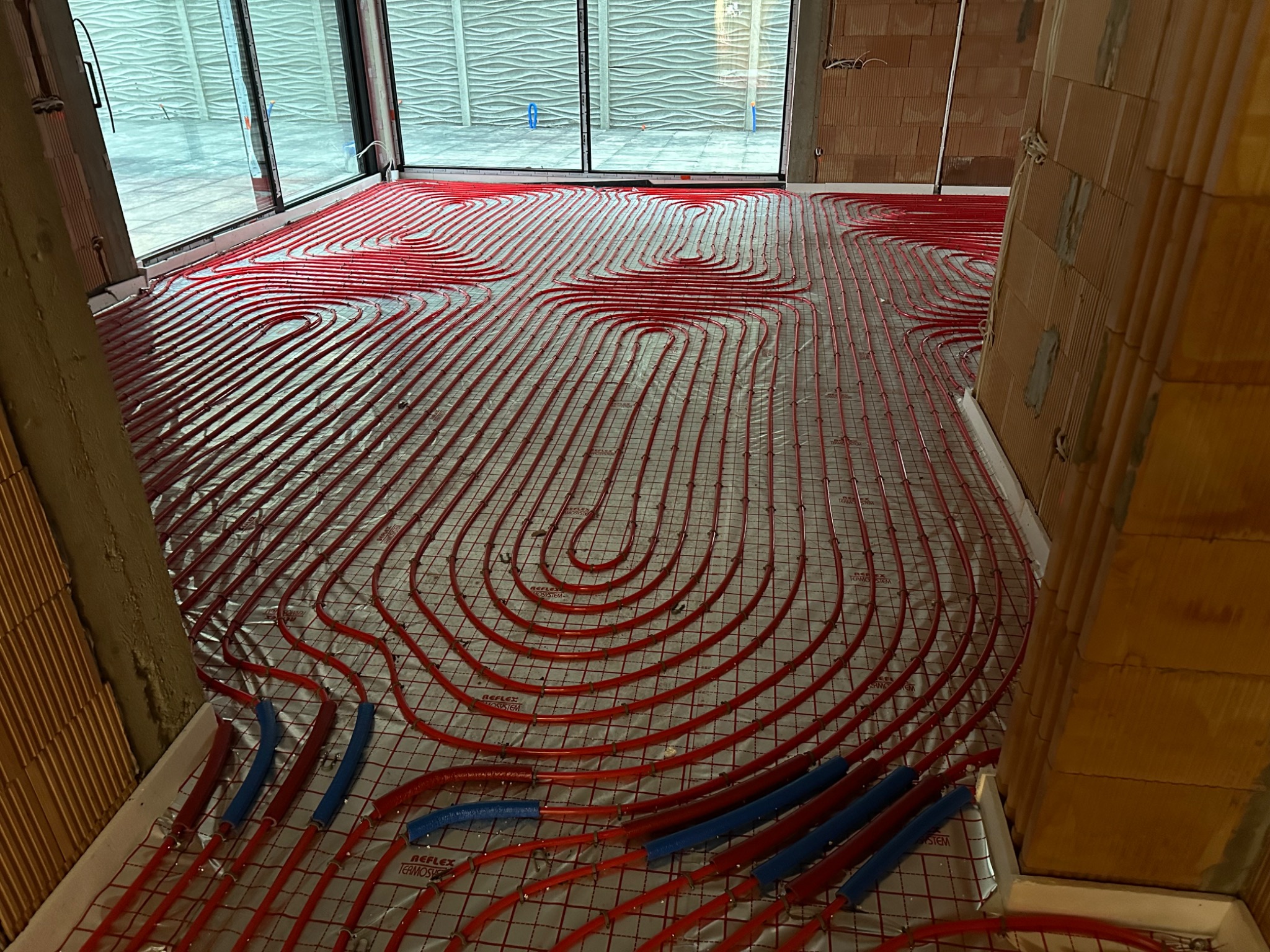 Czerwone rury ogrzewania podłogowego ułożone na folii refleksyjnej w surowym wnętrzu z oknami. Widoczne fragmenty ścian z cegły i niebieskie elementy mocujące rury.
