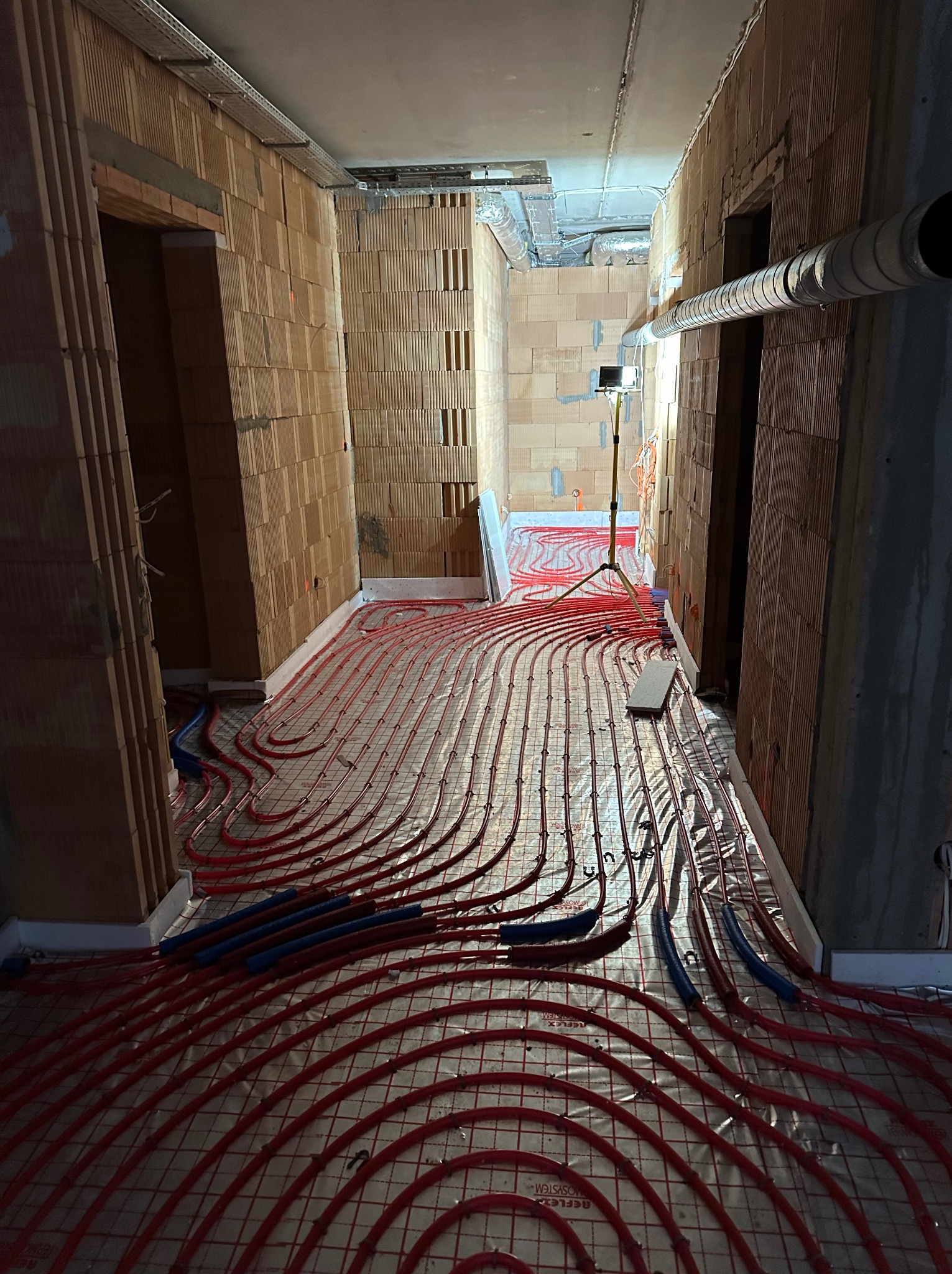 Czerwone rury ogrzewania podłogowego ułożone na siatce w surowym wnętrzu z ceglanymi ścianami. Widoczny fragment instalacji wentylacyjnej pod sufitem i oświetlenie budowlane.