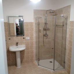 NAR-BUD - Jasna łazienka po remoncie: kabina prysznicowa z chromowaną armaturą, umywalka na postumencie i lustro z odbiciem osoby. Beżowe płytki na ścianach i podłodze.