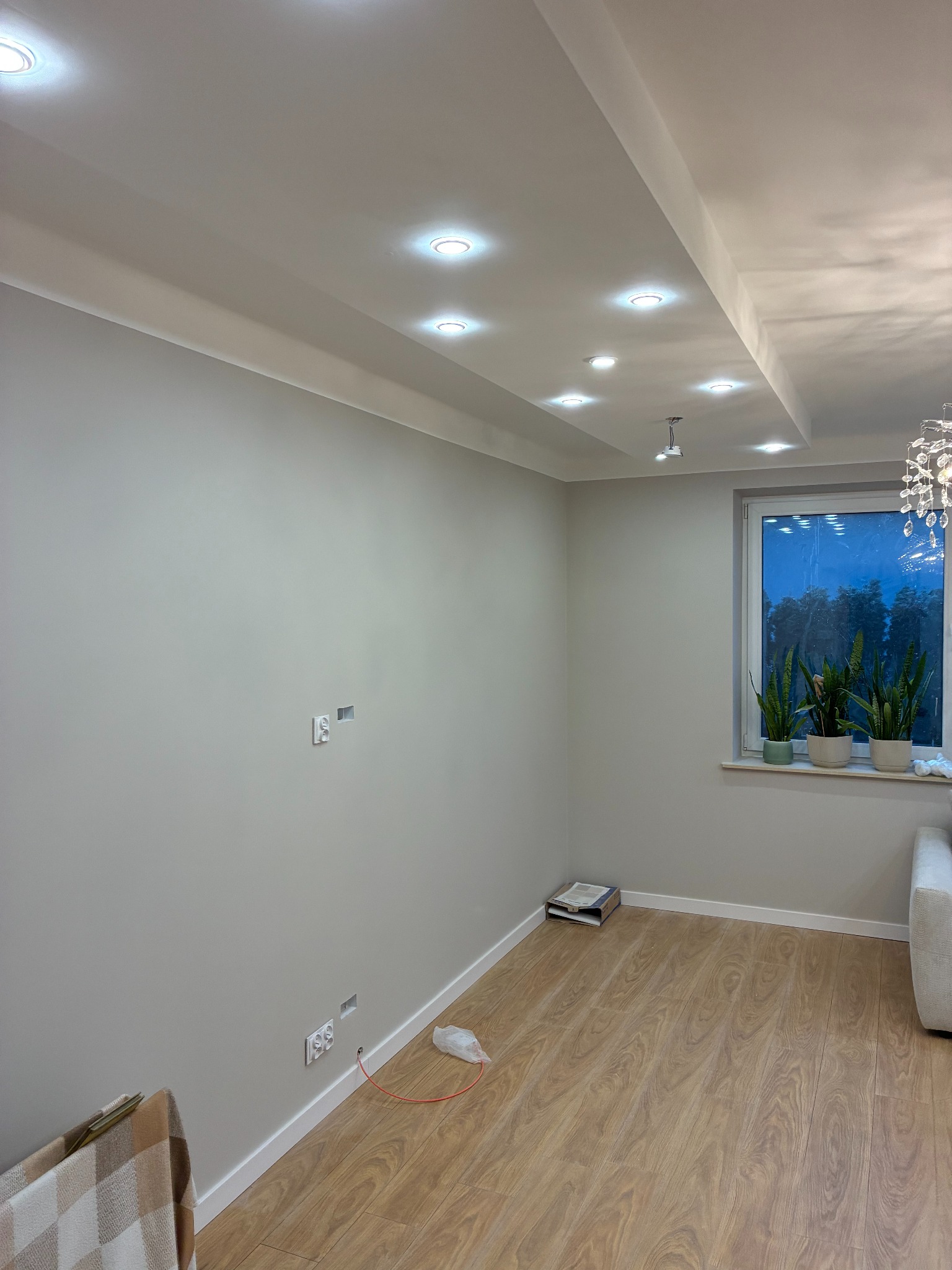 Jasny salon po remoncie: świeżo pomalowane ściany, drewniana podłoga, okno z roślinami i nowoczesne oświetlenie sufitowe. Widoczne gniazdka elektryczne.