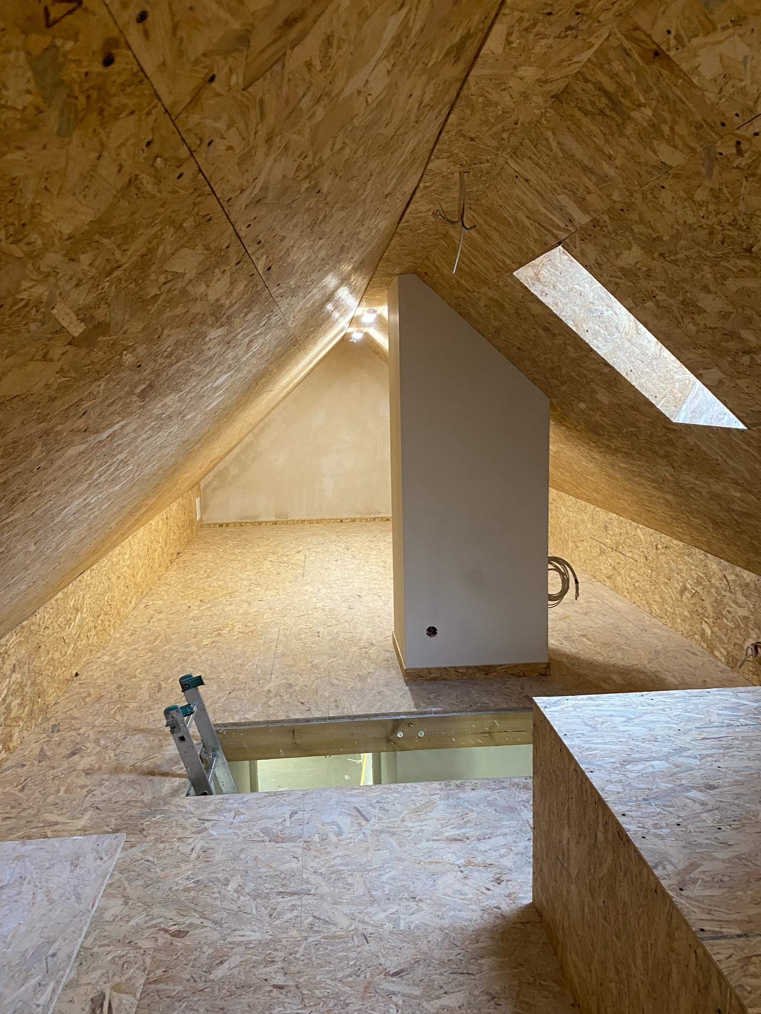 Zabudowa poddasza z płyt OSB, widoczne okno dachowe, fragment schodów i komin wentylacyjny. Surowe wykończenie, etap prac wykończeniowych.