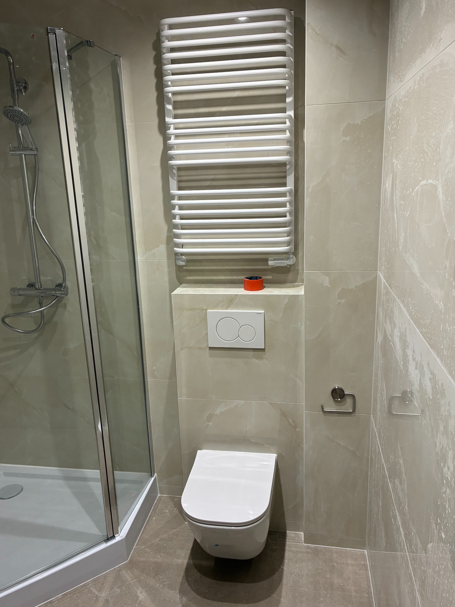 Wykończona łazienka z kabiną prysznicową, grzejnikiem drabinkowym i podwieszaną toaletą. Beżowe płytki imitujące marmur na ścianach i podłodze.