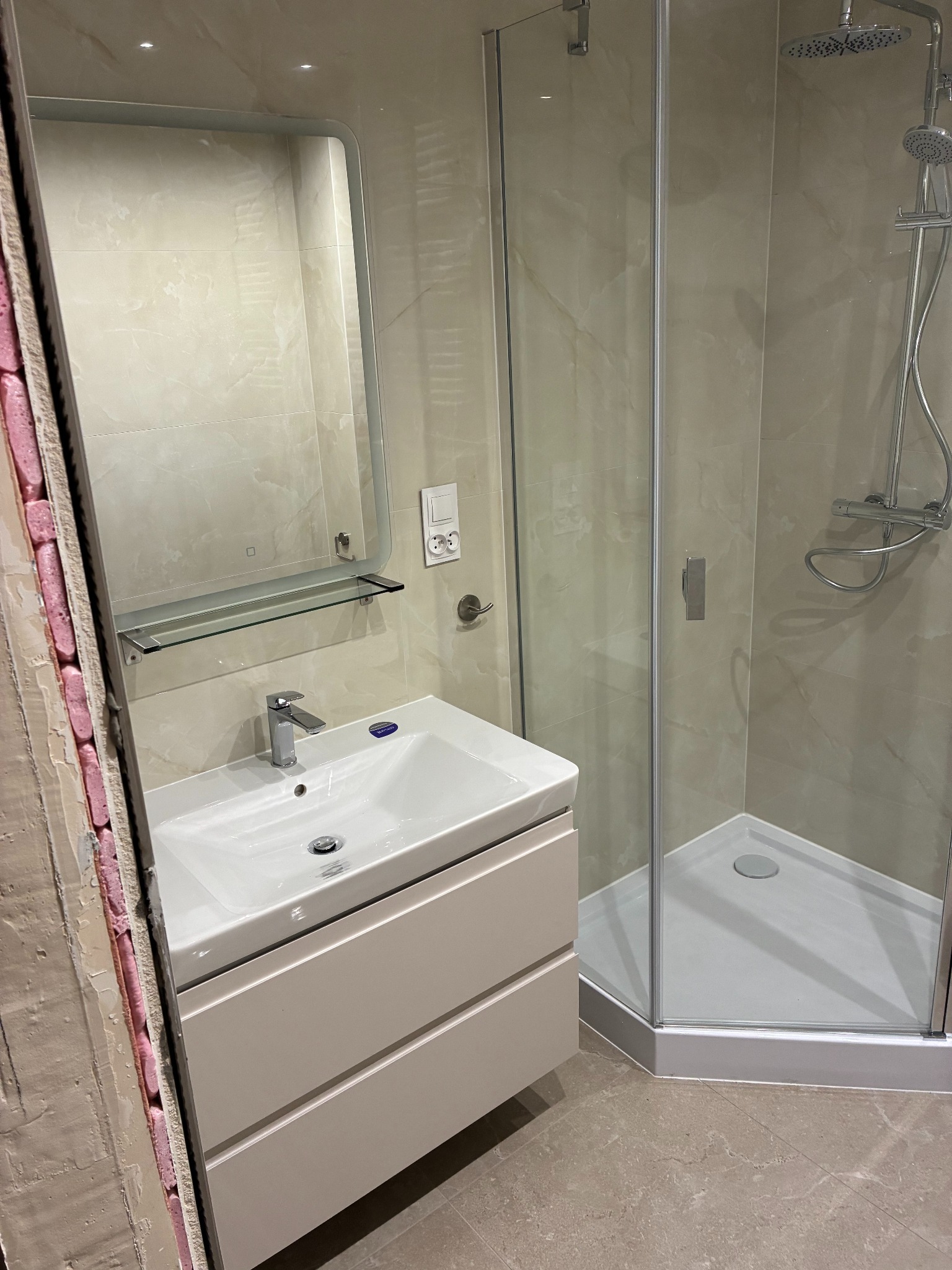 Wykończona łazienka z kabiną prysznicową i umywalką z szafką w beżowej tonacji. Widoczny fragment ściany w trakcie wykończenia.