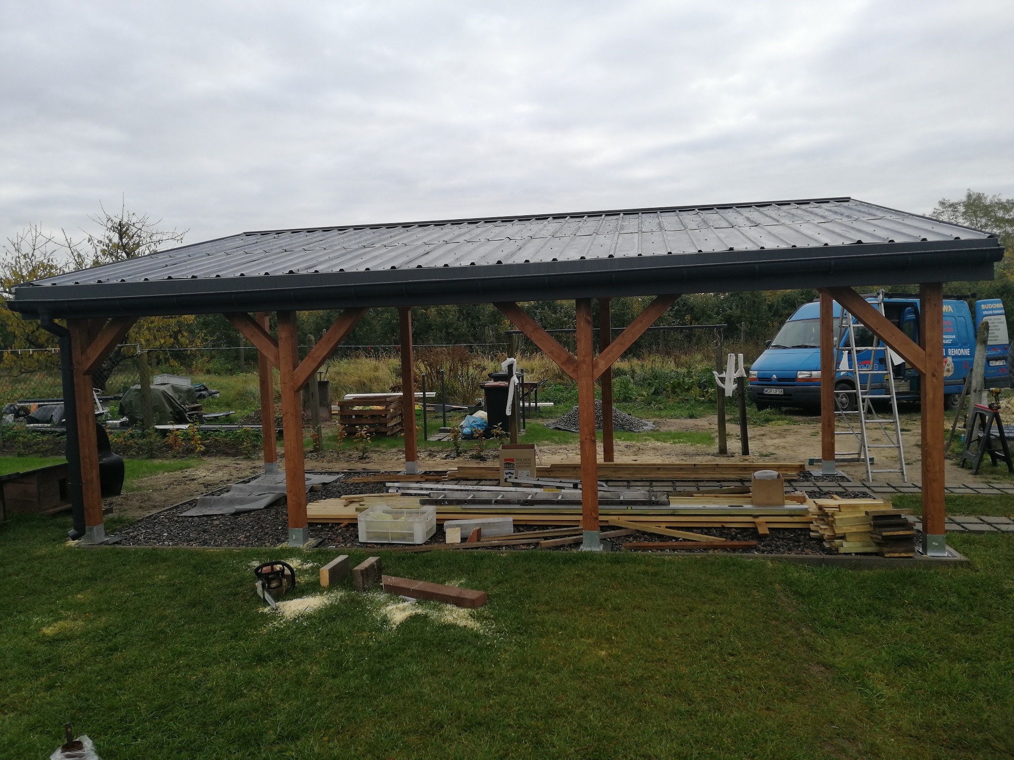 W trakcie budowy wiaty: drewniana konstrukcja z metalowym dachem, widoczne narzędzia i materiały budowlane, na trawie leży piła łańcuchowa, w tle samochód dostawczy.
