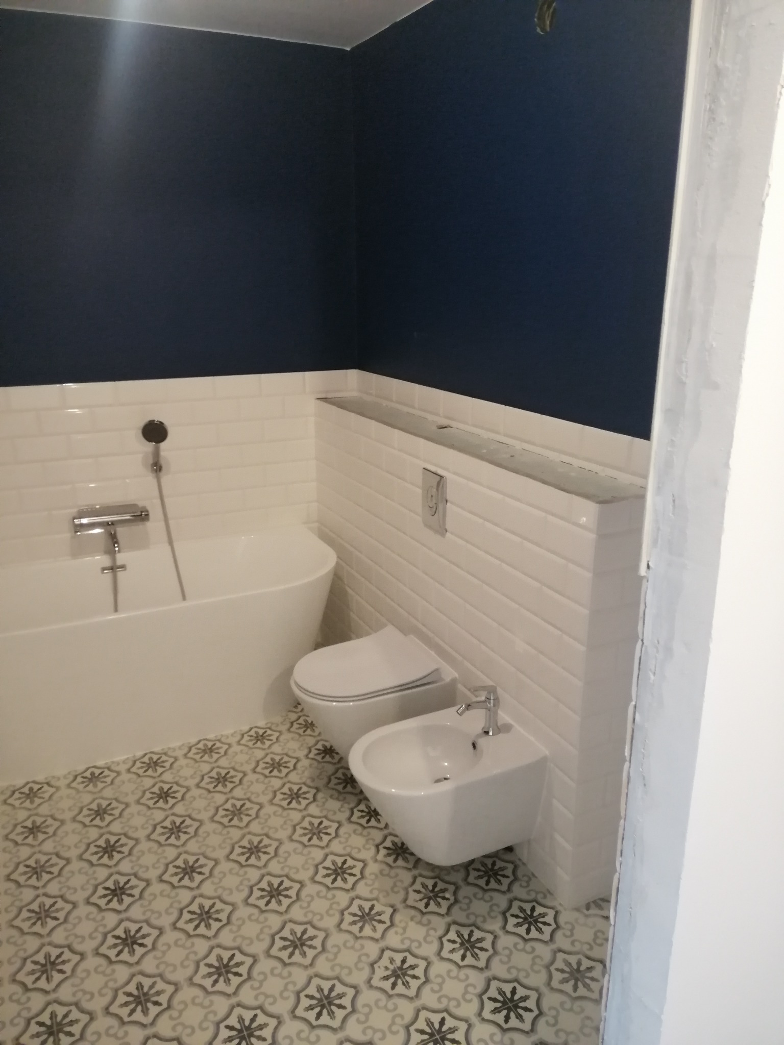 Nowoczesna łazienka z granatową ścianą, białą armaturą (wanna, toaleta, bidet) i wzorzystą podłogą. Widoczne białe płytki typu metro na ścianach.