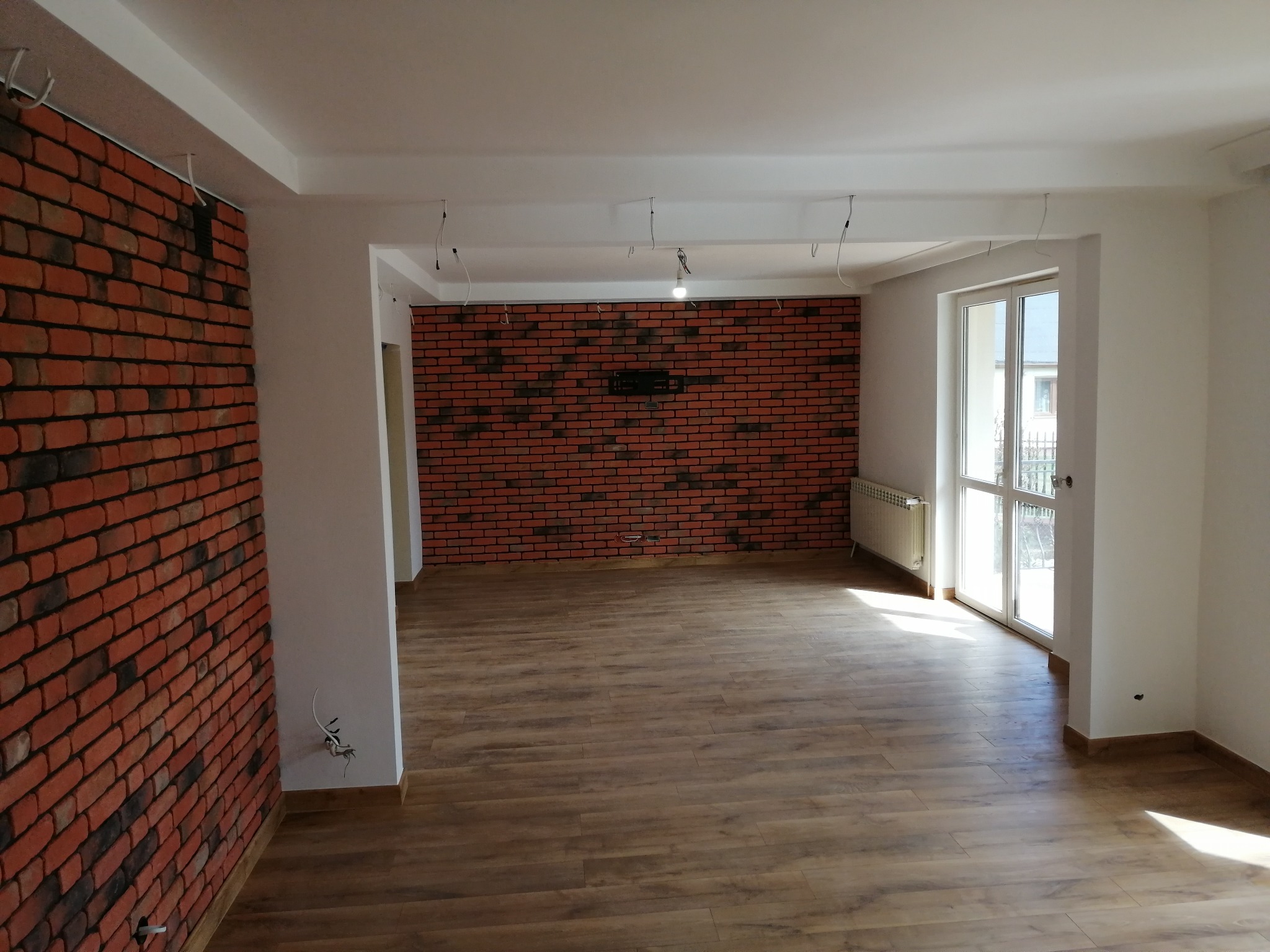 Salon po wykończeniu: drewnopodobna podłoga, ściana z cegły dekoracyjnej, białe ściany i sufit. Widoczne przewody elektryczne. Nowoczesny, minimalistyczny design.