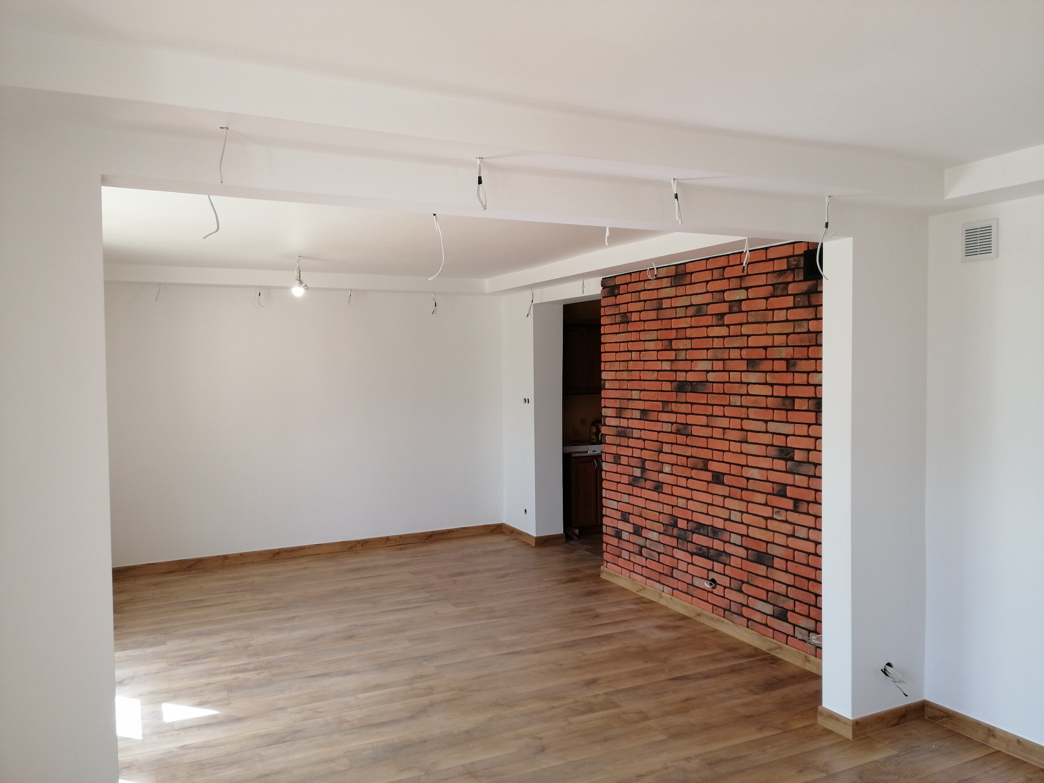 Salon po wykończeniu: drewnopodobna podłoga, białe ściany i fragment ściany z cegły dekoracyjnej. Widoczne wystające kable oświetleniowe.