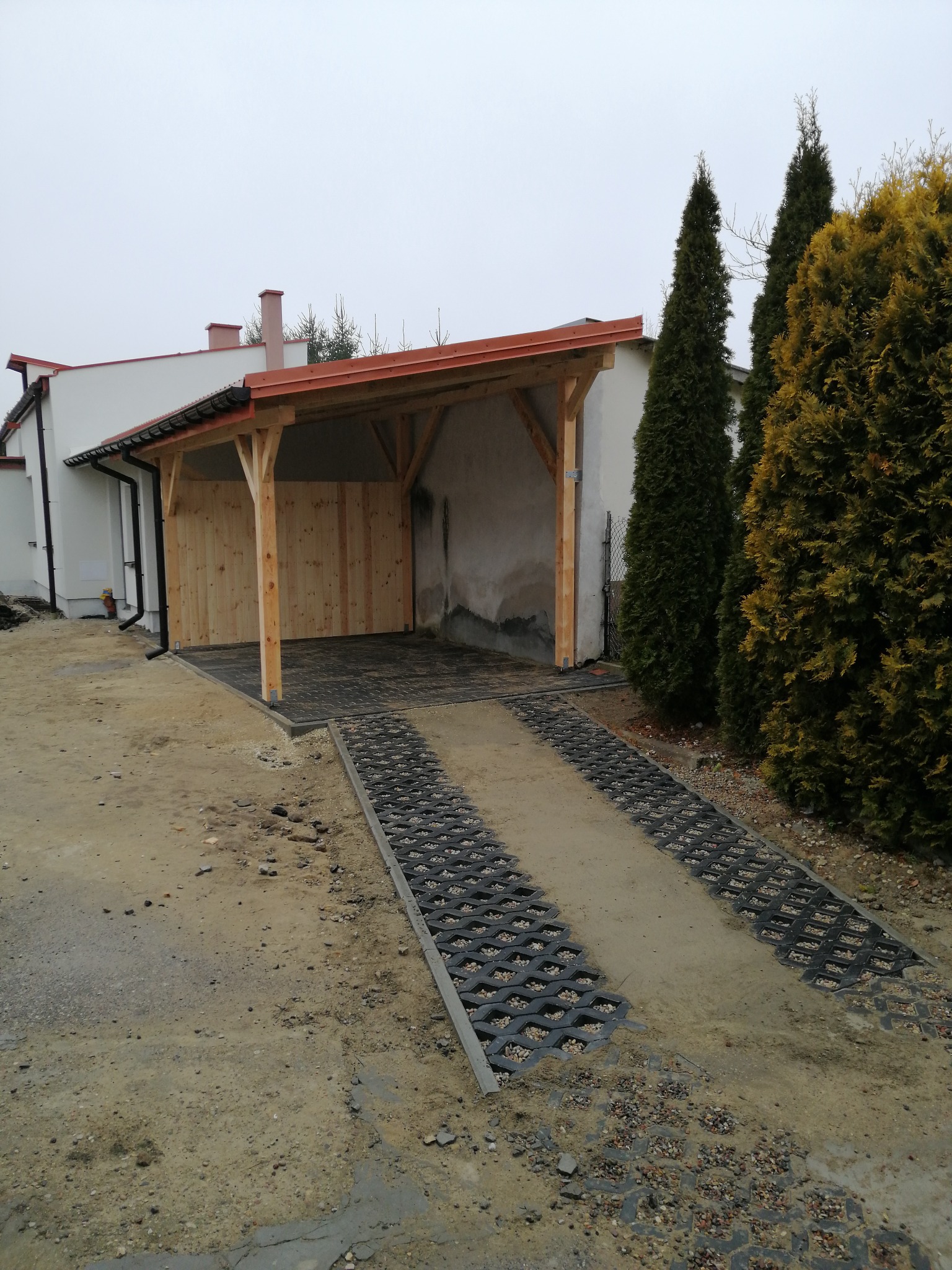 Drewniana wiata przylegająca do budynku, z wyłożonym podjazdem z ażurowych płyt. Widoczne elementy konstrukcyjne i częściowo obudowane ściany. Tło: budynek i zieleń.