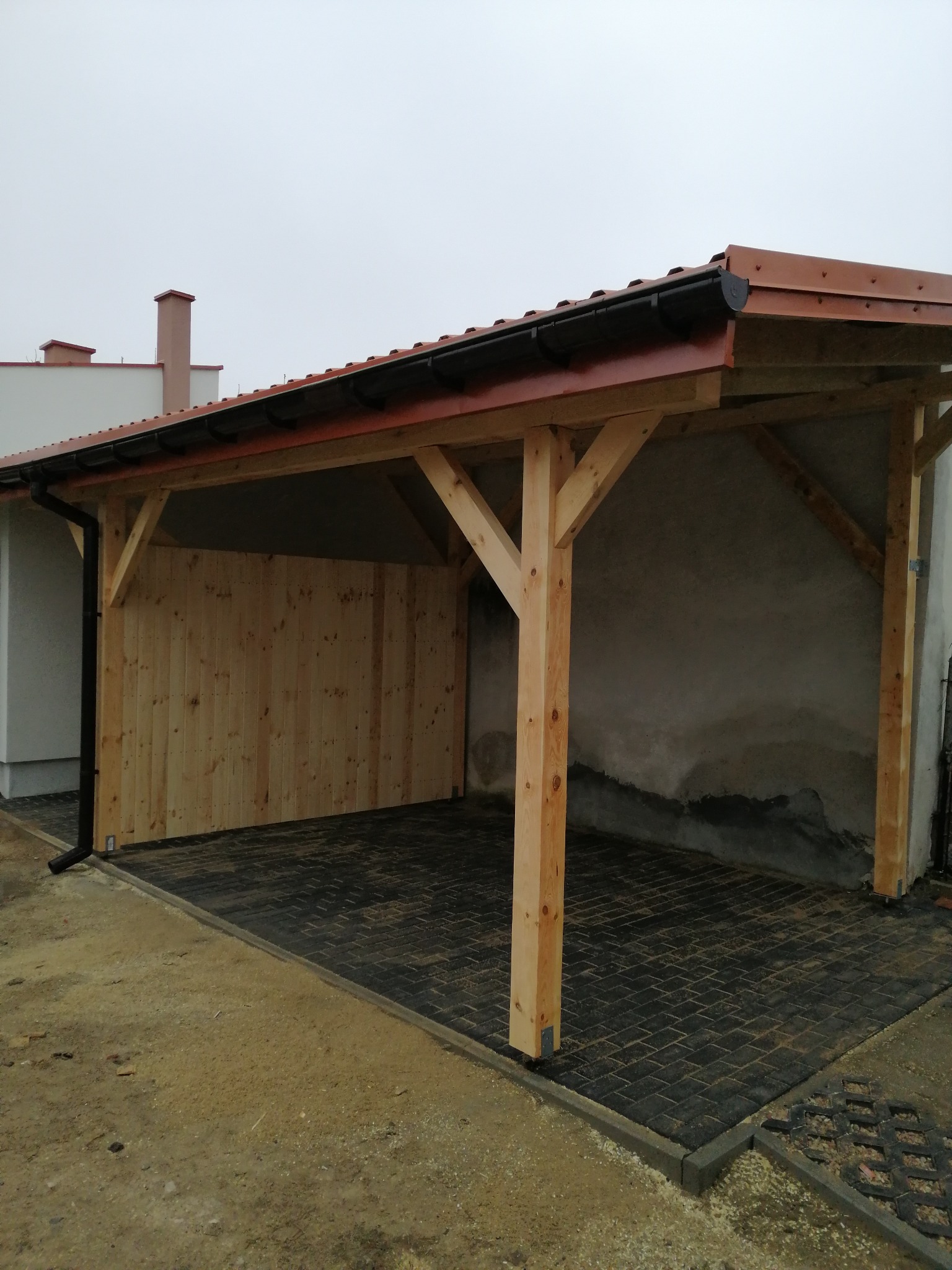 Drewniana wiata z brązowym dachem i czarnymi rynnami, częściowo obita deskami, na posesji z kostką brukową. Widok z zewnątrz, pochmurne niebo w tle.