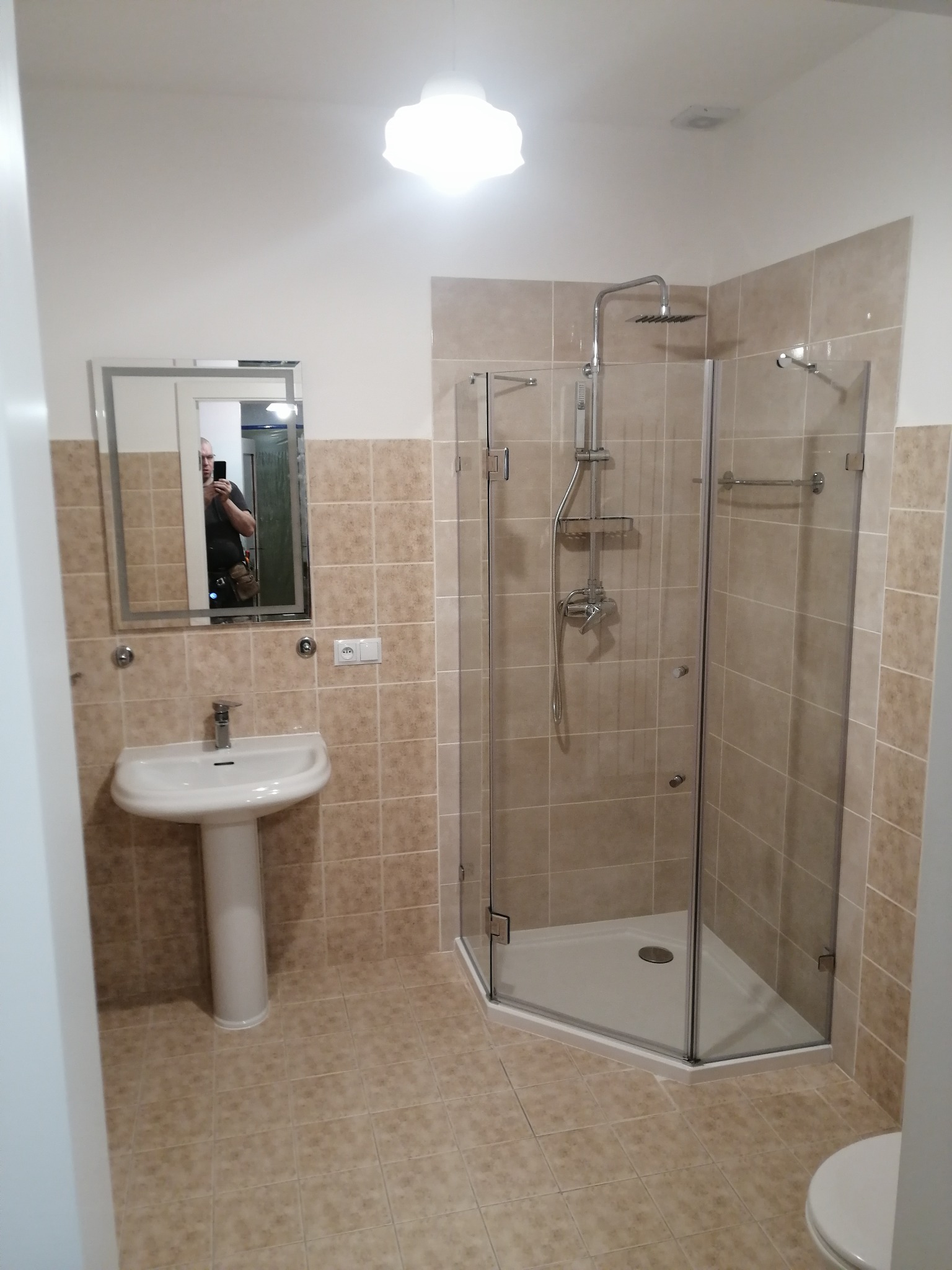 Jasna łazienka po remoncie: kabina prysznicowa z chromowaną armaturą, umywalka na postumencie i lustro z odbiciem osoby. Beżowe płytki na ścianach i podłodze.