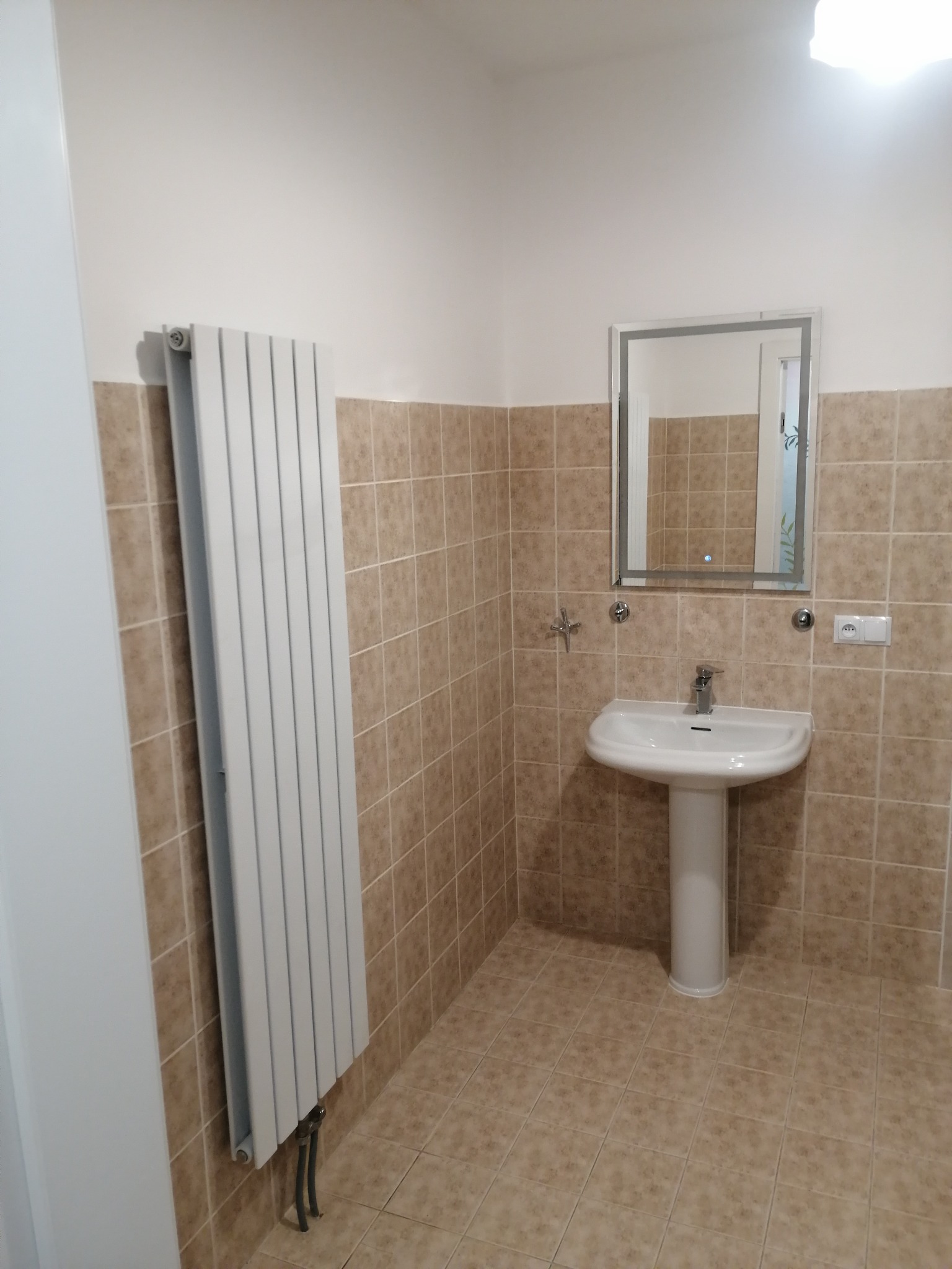 Wykończona łazienka z beżowymi płytkami, białą umywalką na postumencie, grzejnikiem żeberkowym i lustrem. Widoczne detale: bateria, włącznik, zawór.