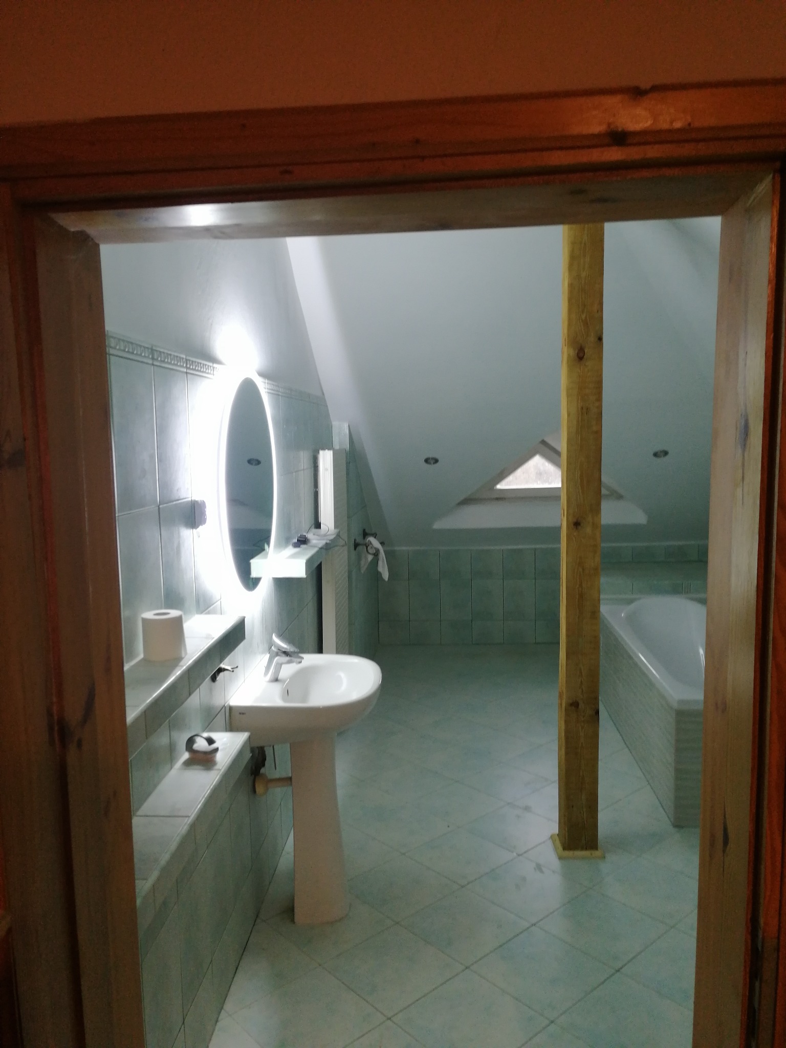 Łazienka na poddaszu z owalnym lustrem z podświetleniem LED, białą umywalką na postumencie i wanną. Widok przez drewniane futryny drzwi.