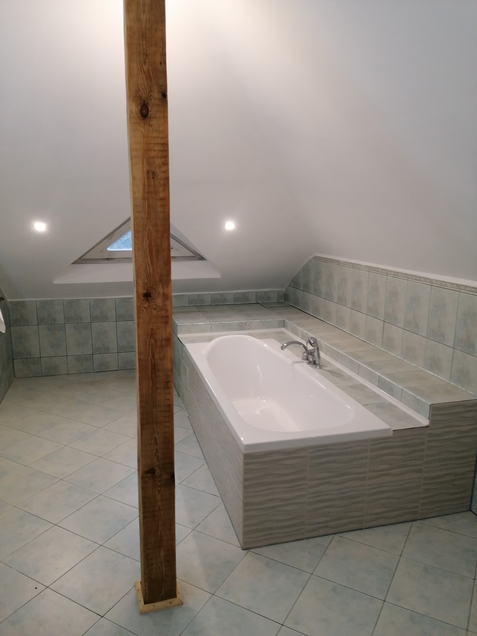 Wykończona łazienka na poddaszu z wanną obudowaną płytkami i drewnianym słupem konstrukcyjnym. Ukośny sufit z oknem. Płytki podłogowe i ścienne w odcieniach szarości.