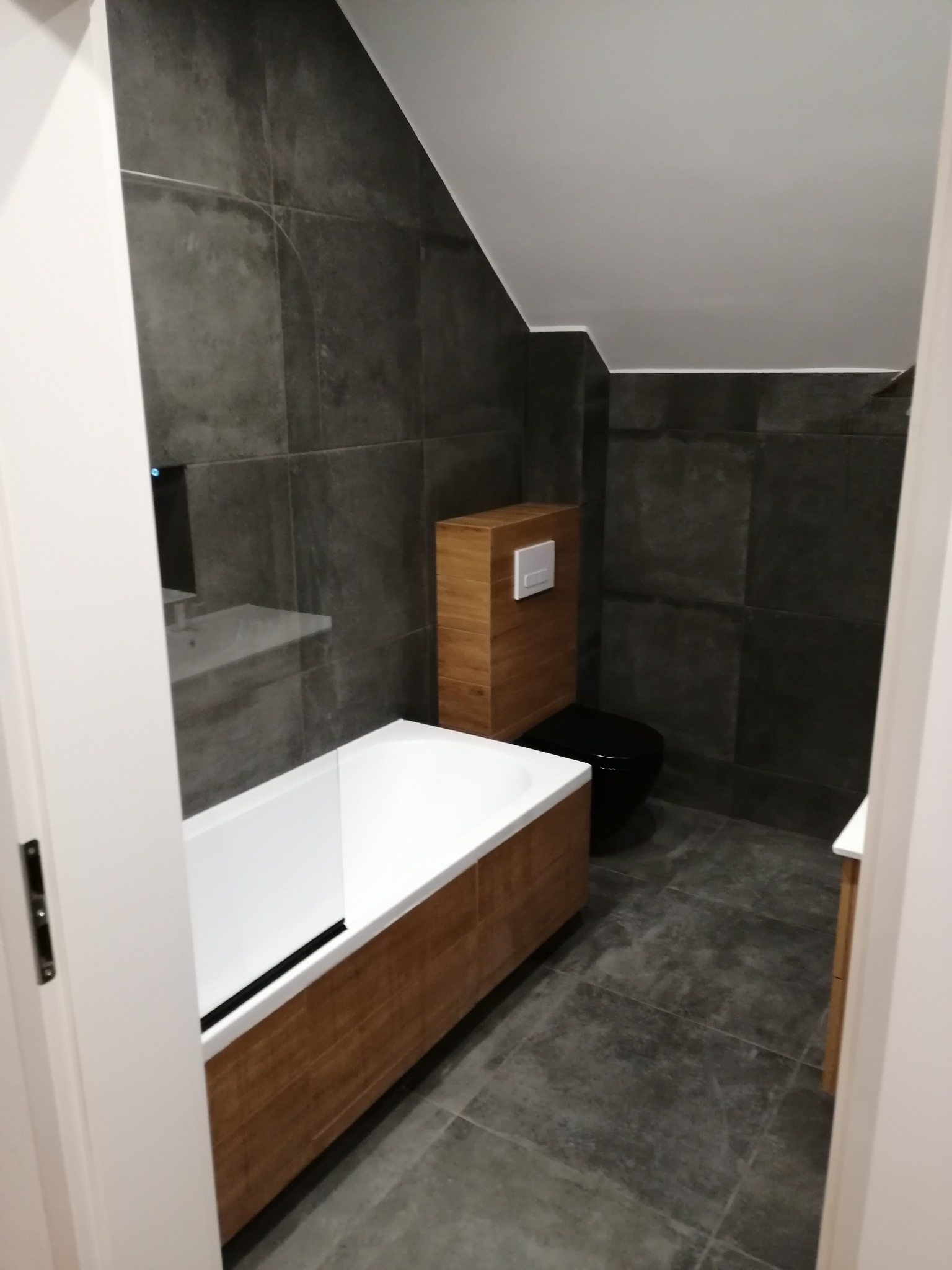 Nowoczesna łazienka na poddaszu z szarymi płytkami, drewnianą obudową wanny i toaletą w kolorze czarnym. Widok zza otwartych, białych drzwi.