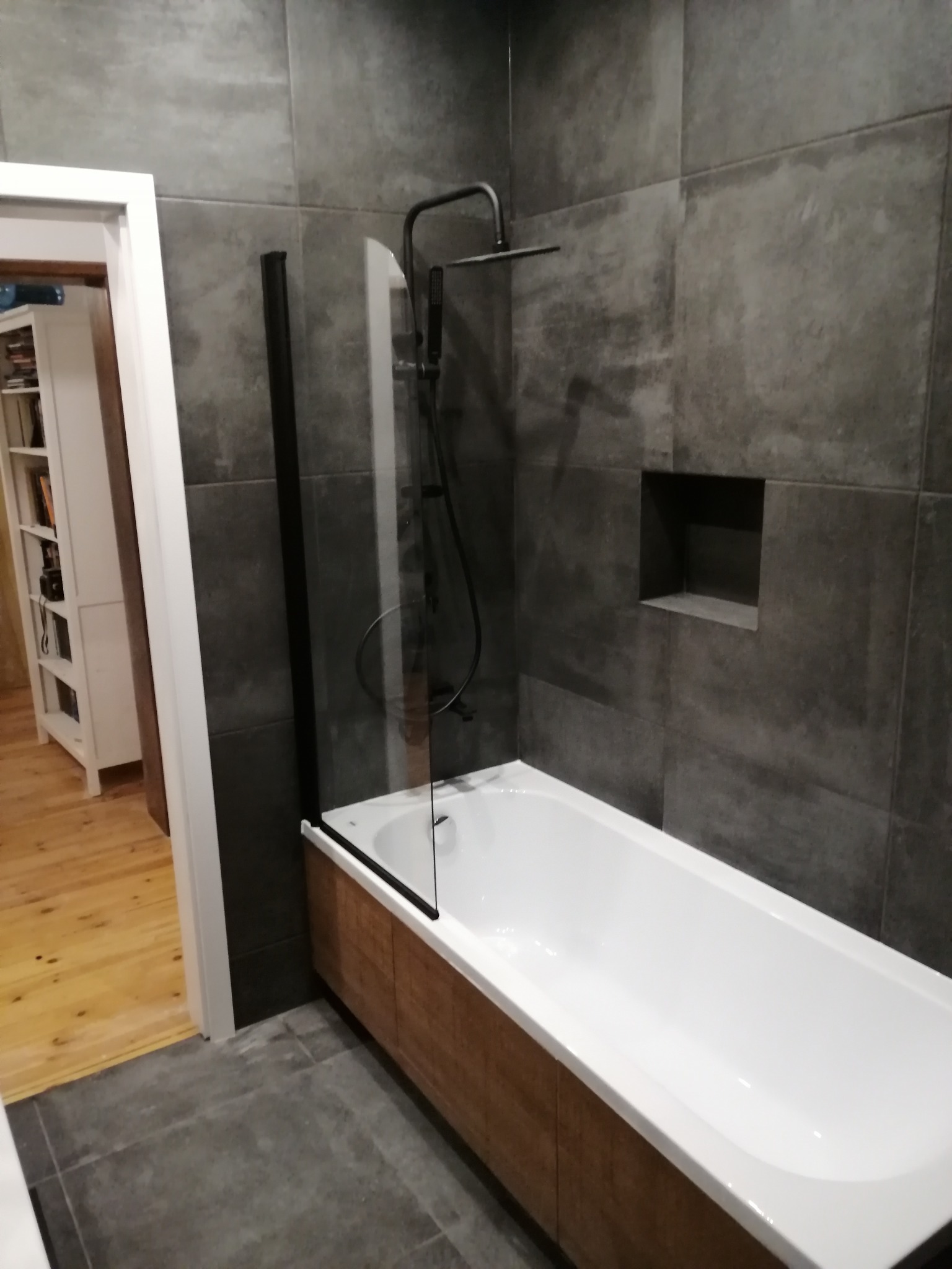 Nowoczesna łazienka z wanną obudowaną drewnem, szarą glazurą i czarną armaturą. Kabina prysznicowa częściowo szklana z czarnymi profilami. W tle widoczne otwarte drzwi.