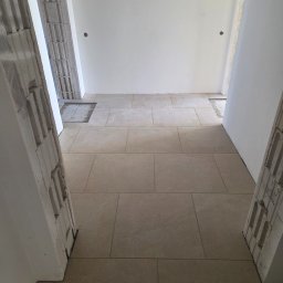 ArtBud - Ułożona, beżowa podłoga z płytek w korytarzu, widok z góry. Fragmenty ścian z pustaków i gładzią. Widoczne niedokończone prace przy podłodze.