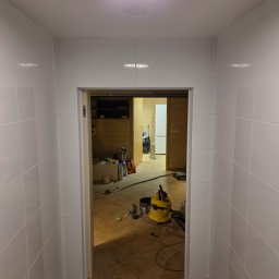 ArtBud - Białe, kwadratowe płytki na ścianach korytarza prowadzącego do remontowanego pomieszczenia. Widoczny odkurzacz przemysłowy i materiały budowlane.