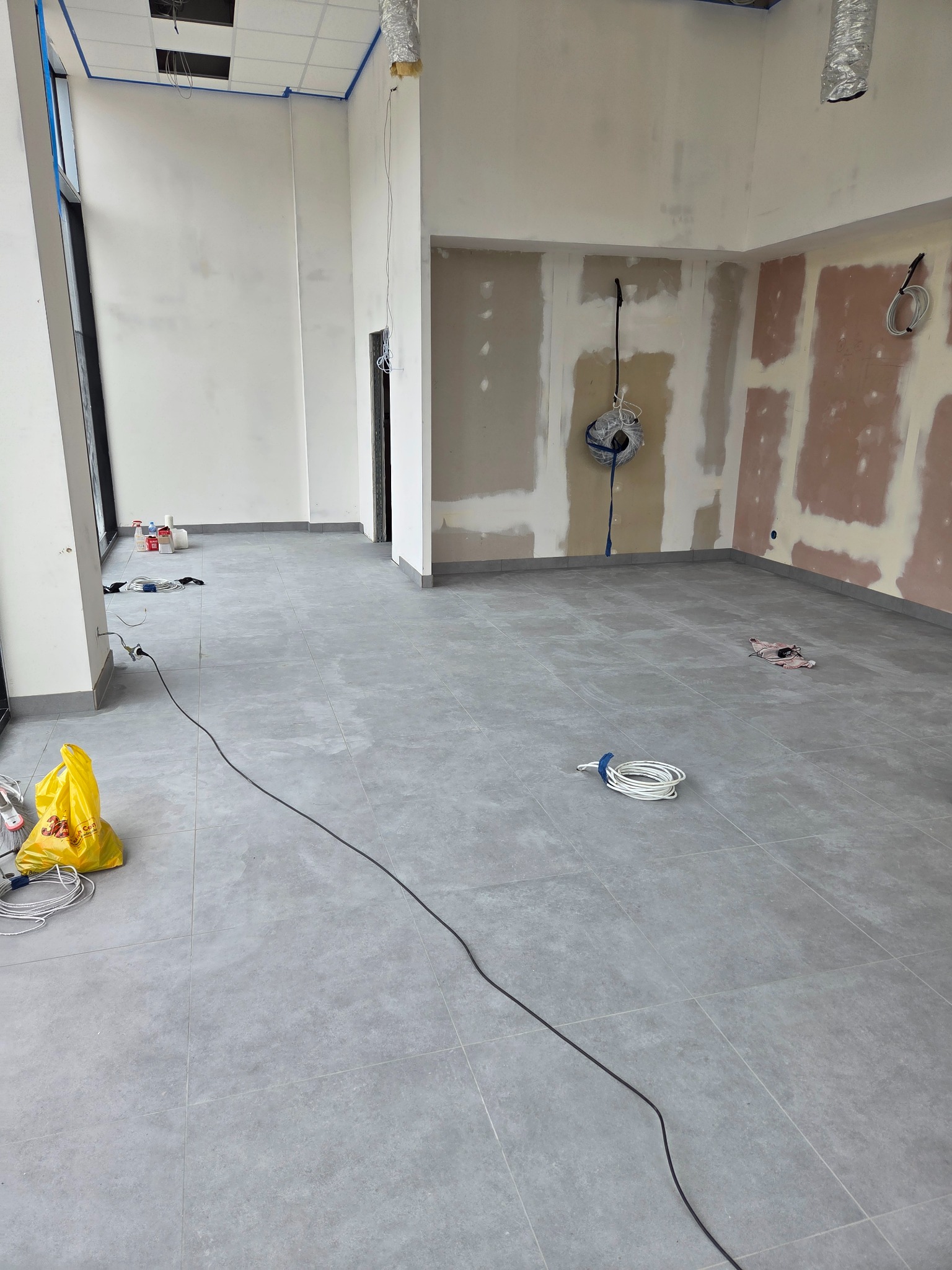 Wnętrze w trakcie wykończenia: szare płytki podłogowe, ściany z fragmentami płyt gips-karton, widoczne przewody i materiały budowlane, taśma malarska przy suficie.