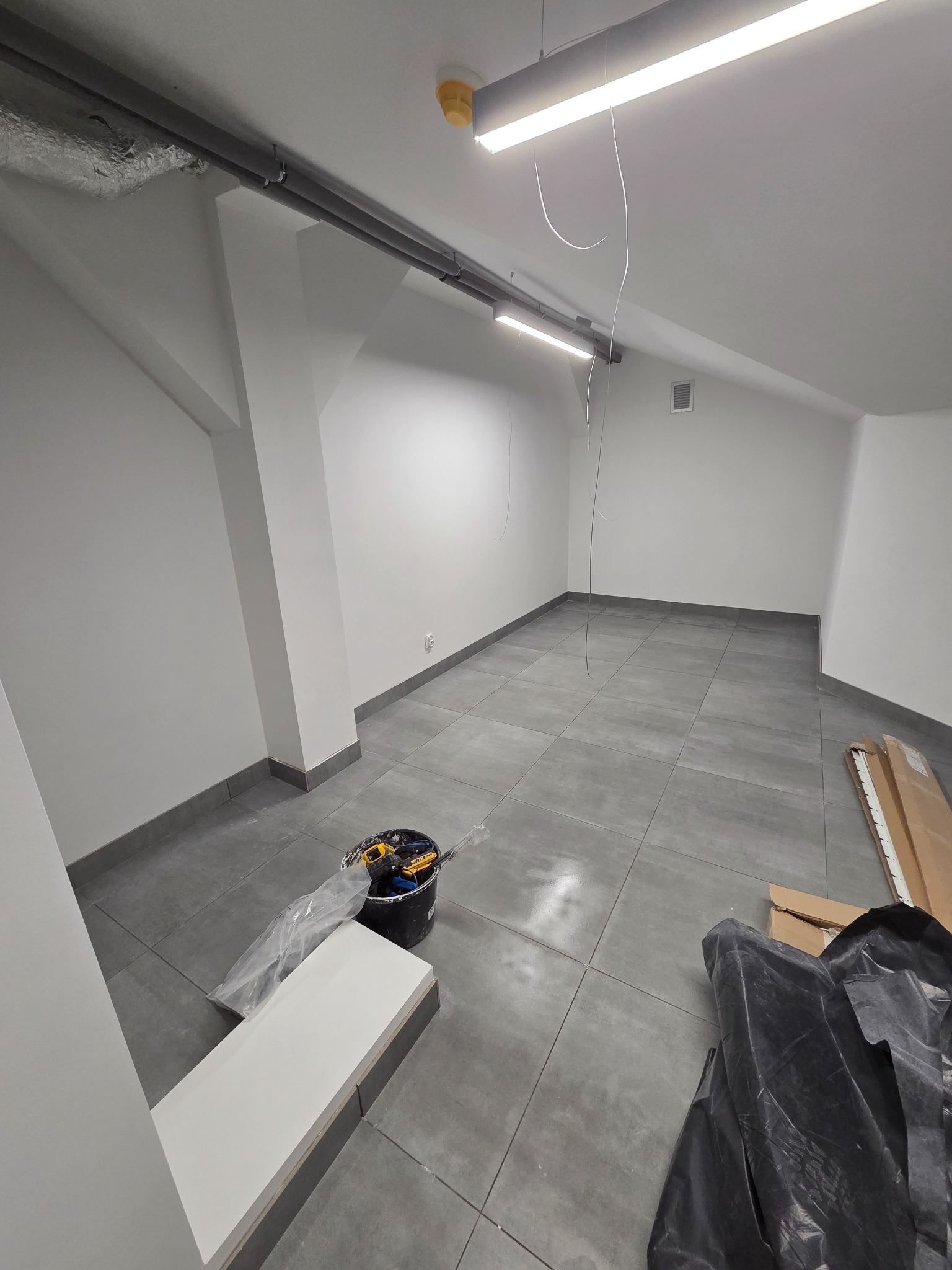 Wykończone wnętrze z szarymi płytkami podłogowymi i świeżo pomalowanymi ścianami. Widoczne narzędzia i materiały budowlane, sugerujące prace wykończeniowe w toku.