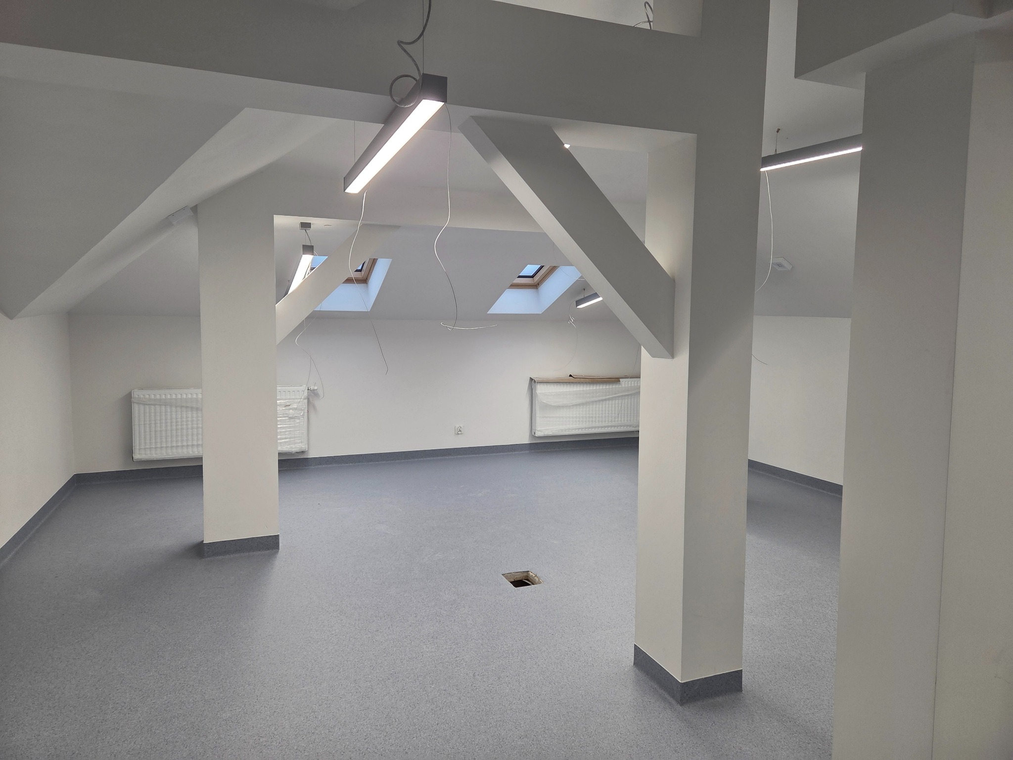 Jasne, przestronne poddasze po remoncie z szarą podłogą, białymi ścianami i kolumnami. Widoczne okna dachowe i nowoczesne oświetlenie.