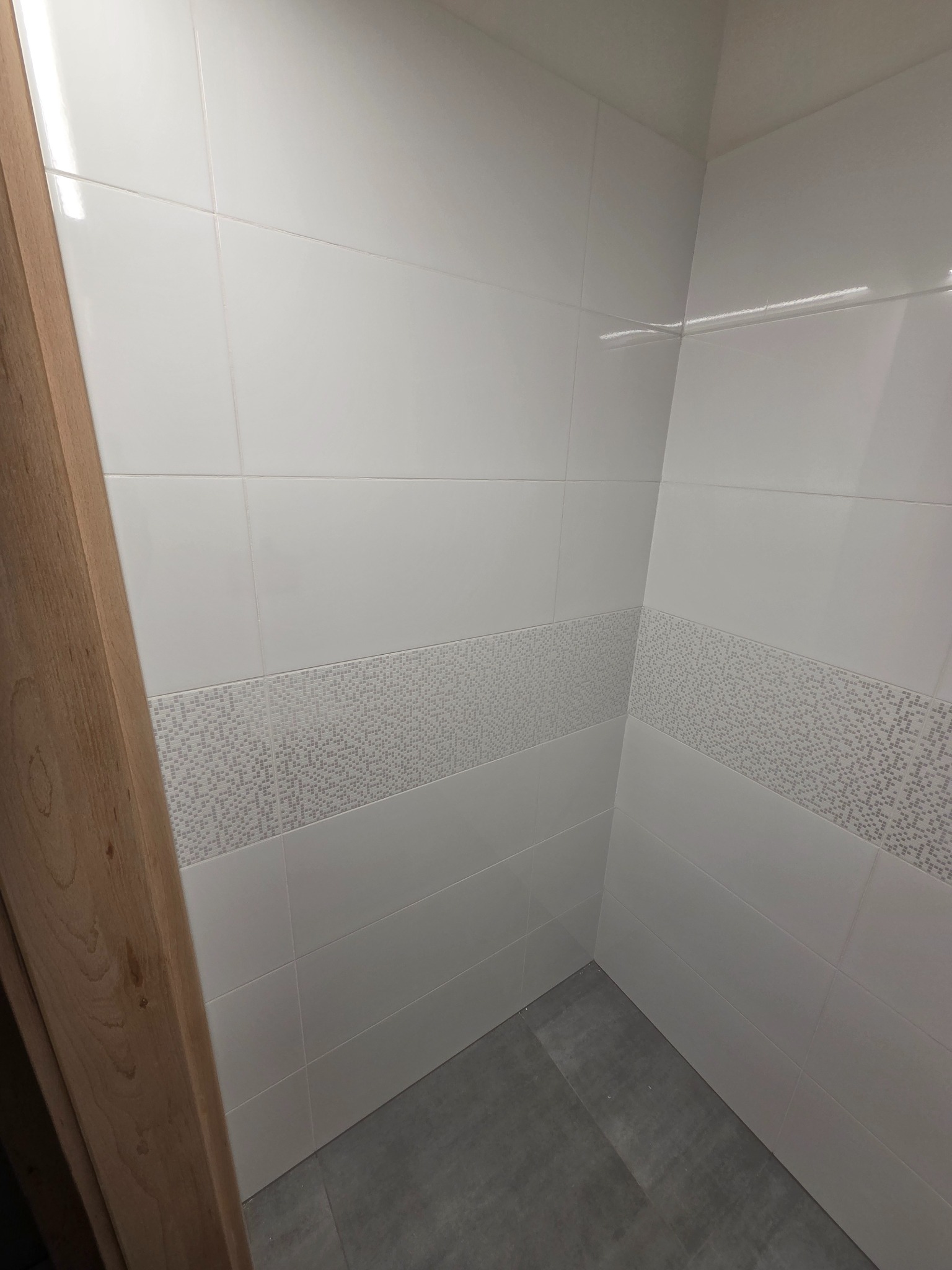 Wykończona łazienka z białymi płytkami i szarymi akcentami mozaiki. Widoczny fragment drewnianej ościeżnicy drzwi. Minimalistyczny design, czyste linie.