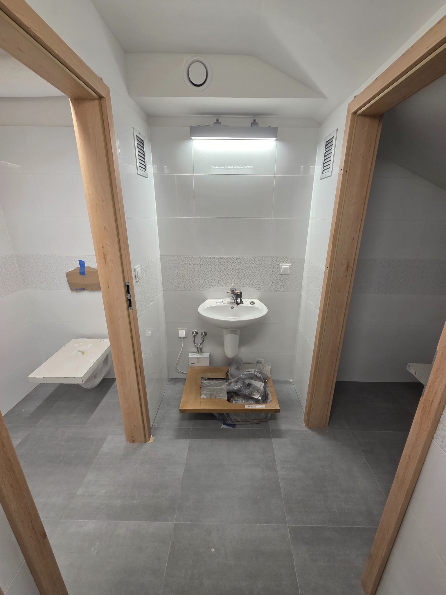 Wnętrze łazienki w trakcie remontu: białe płytki, szara podłoga, umywalka z armaturą, częściowo zamontowane drewniane ościeżnice drzwiowe po obu stronach kadru.