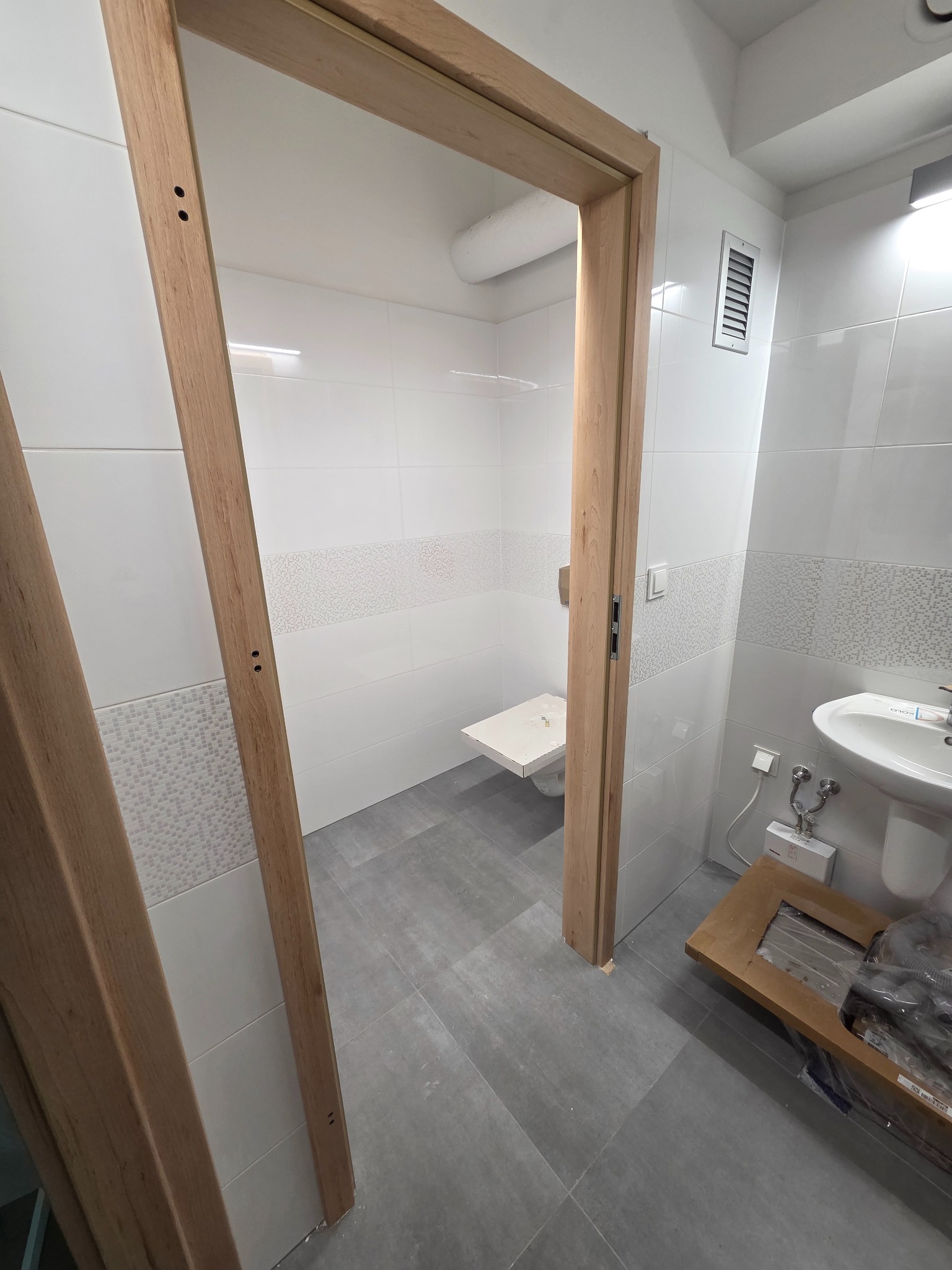 Jasna łazienka z szarymi płytkami podłogowymi, widok przez drewnianą ościeżnicę. Częściowo widoczna umywalka i toaleta. Na podłodze karton z elementami drzwi.