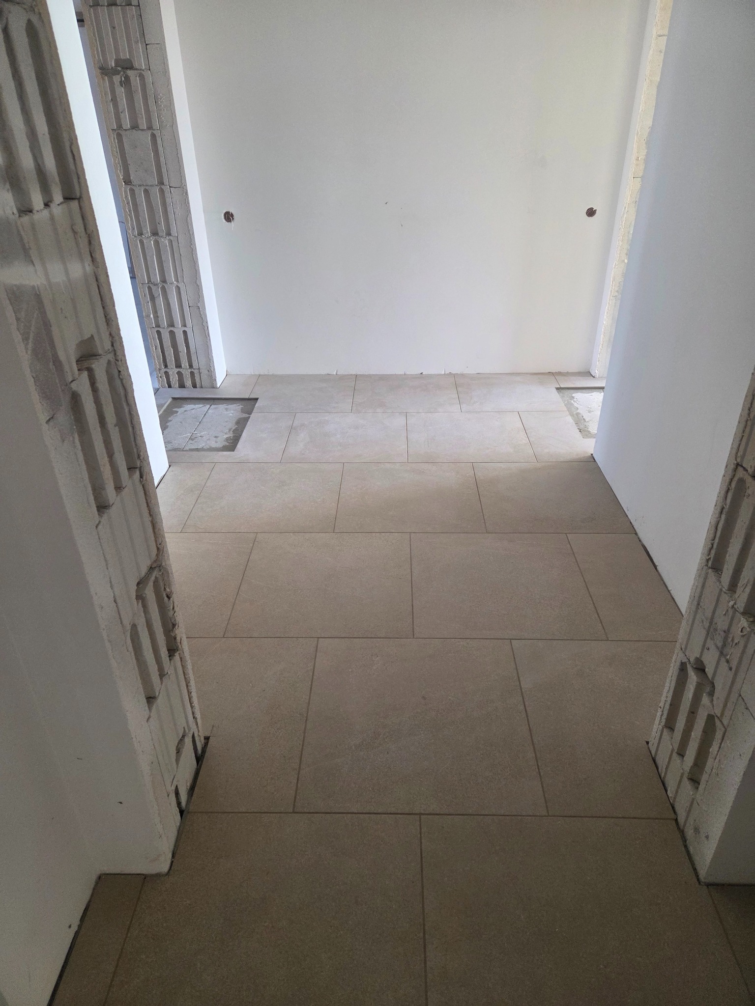 Ułożona, beżowa podłoga z płytek w korytarzu, widok z góry. Fragmenty ścian z pustaków i gładzią. Widoczne niedokończone prace przy podłodze.