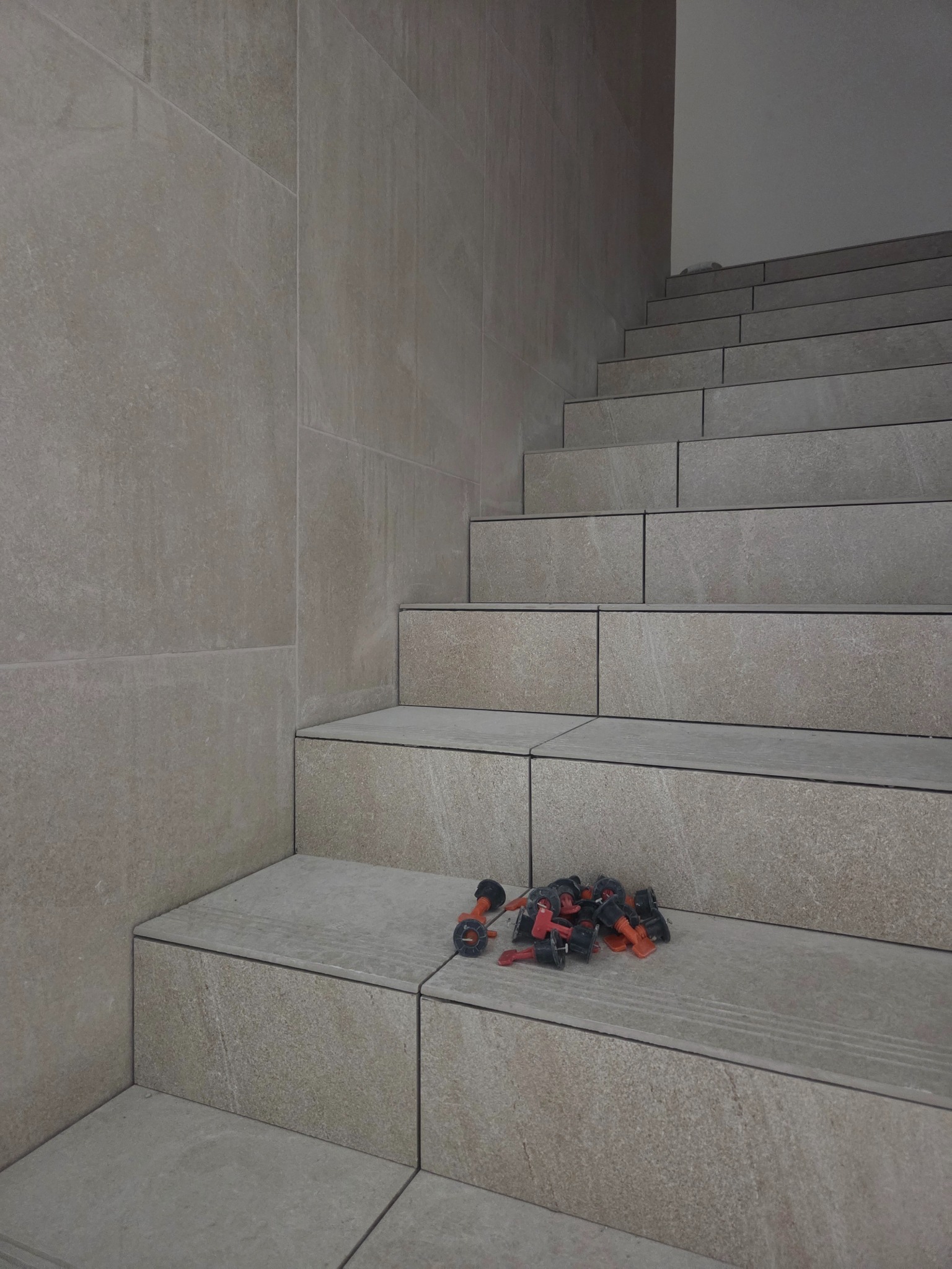 Wykończone schody z płytek w odcieniach beżu i szarości, z widocznymi elementami systemu poziomowania płytek na jednym ze stopni. Minimalistyczny design.