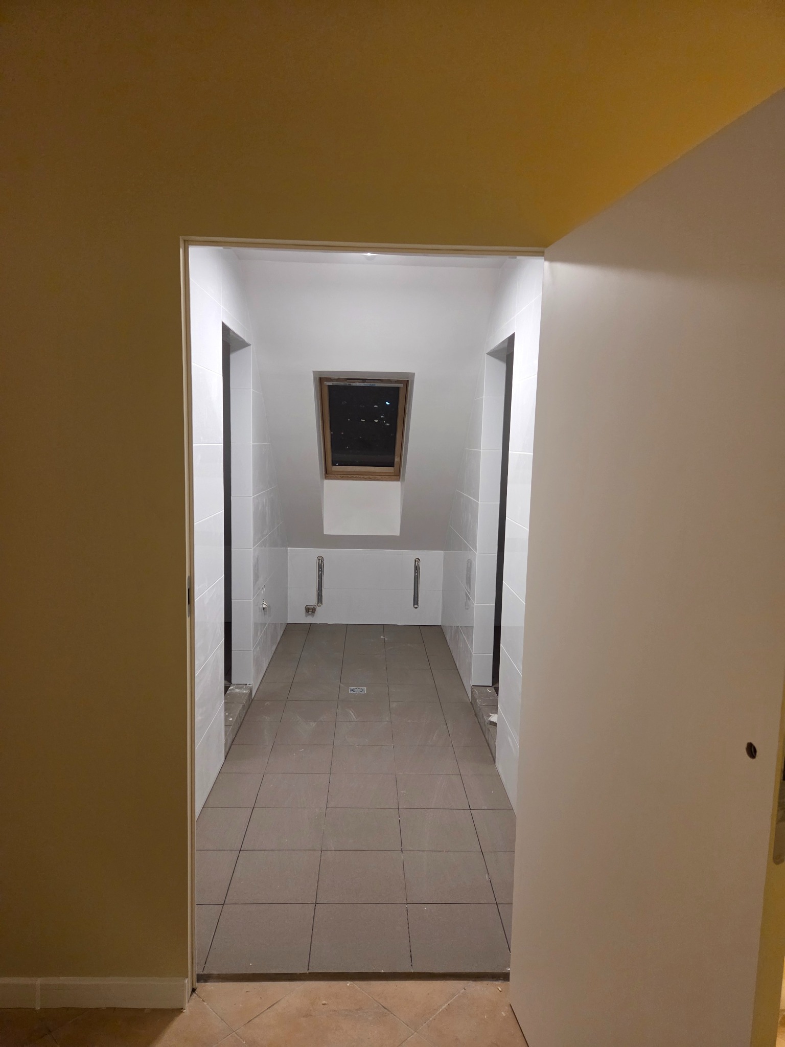 Wykończona łazienka na poddaszu w Szczecinie. Ułożone płytki podłogowe i ścienne, widoczne rury. Ujęcie z korytarza przez otwarte drzwi.