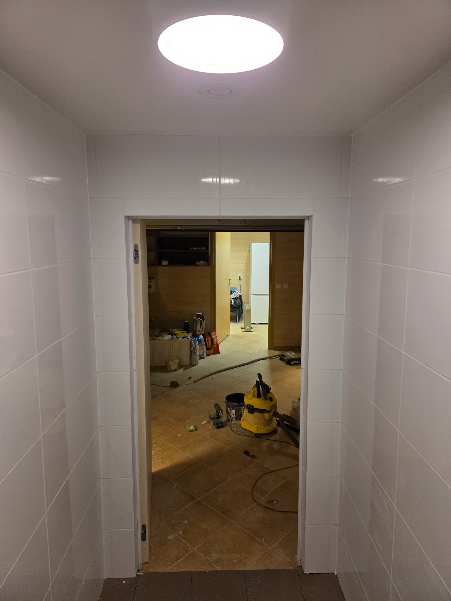 Białe, kwadratowe płytki na ścianach korytarza prowadzącego do remontowanego pomieszczenia. Widoczny odkurzacz przemysłowy i materiały budowlane.