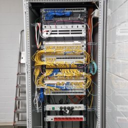 TEHS Netzwerk Solutions Sp. z o.o. - Szafa rack z patch panelem i okablowaniem sieciowym w kolorach niebieskim i żółtym. Widoczna drabina aluminiowa obok szafy.