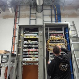 TEHS Netzwerk Solutions Sp. z o.o. - Pracownik w czarnej bluzie konfiguruje kable w szafie rack z urządzeniami sieciowymi. Widoczne kable, przełączniki, drabina i instalacje pod sufitem.
