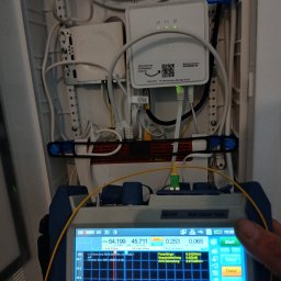 TEHS Netzwerk Solutions Sp. z o.o. - Pomiar światłowodu w szafce telekomunikacyjnej za pomocą testera optycznego AE3100. Widoczne kable, router i wyniki pomiarów na ekranie urządzenia.