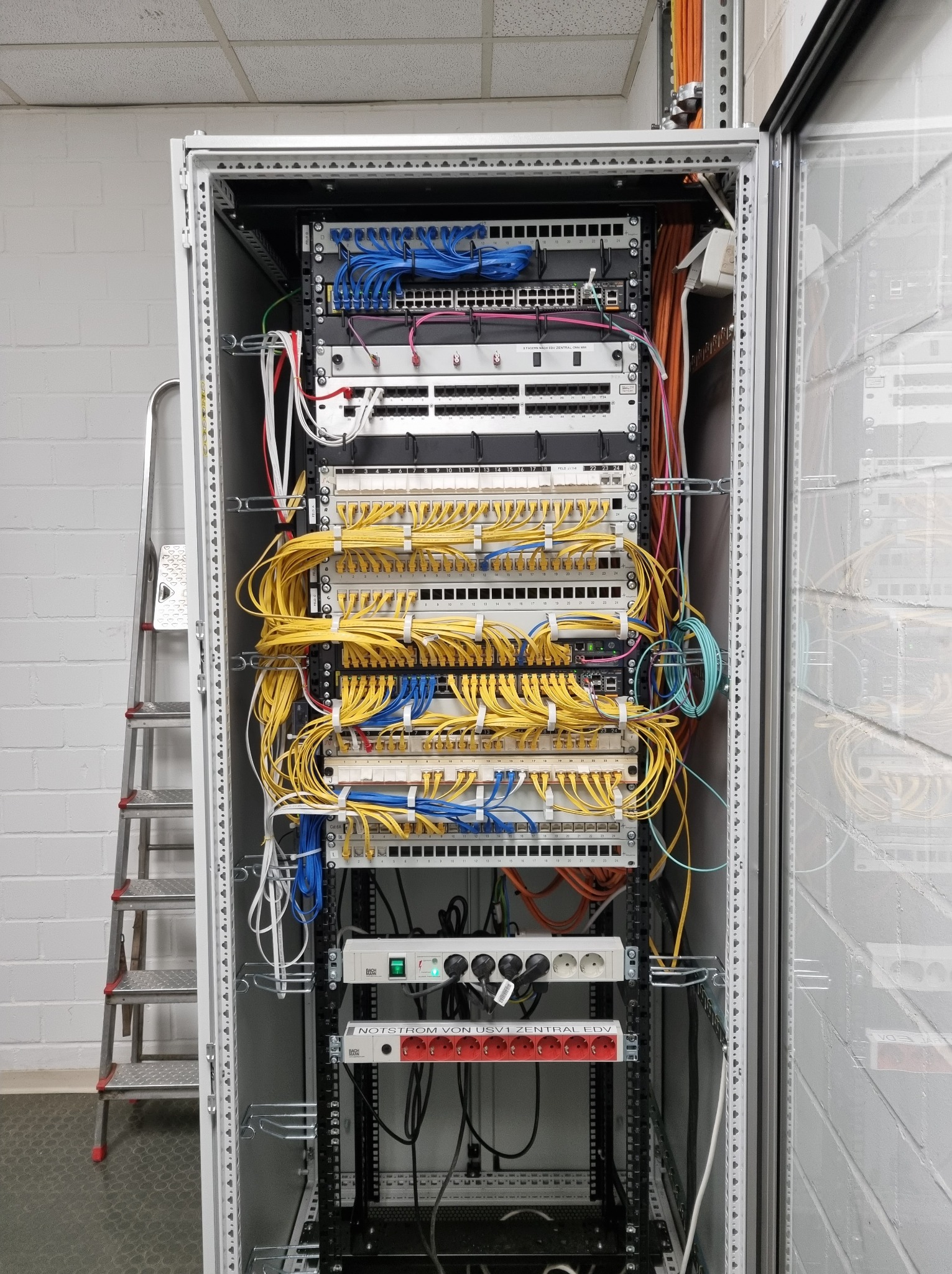 Szafa rack z patch panelem i okablowaniem sieciowym w kolorach niebieskim i żółtym. Widoczna drabina aluminiowa obok szafy.