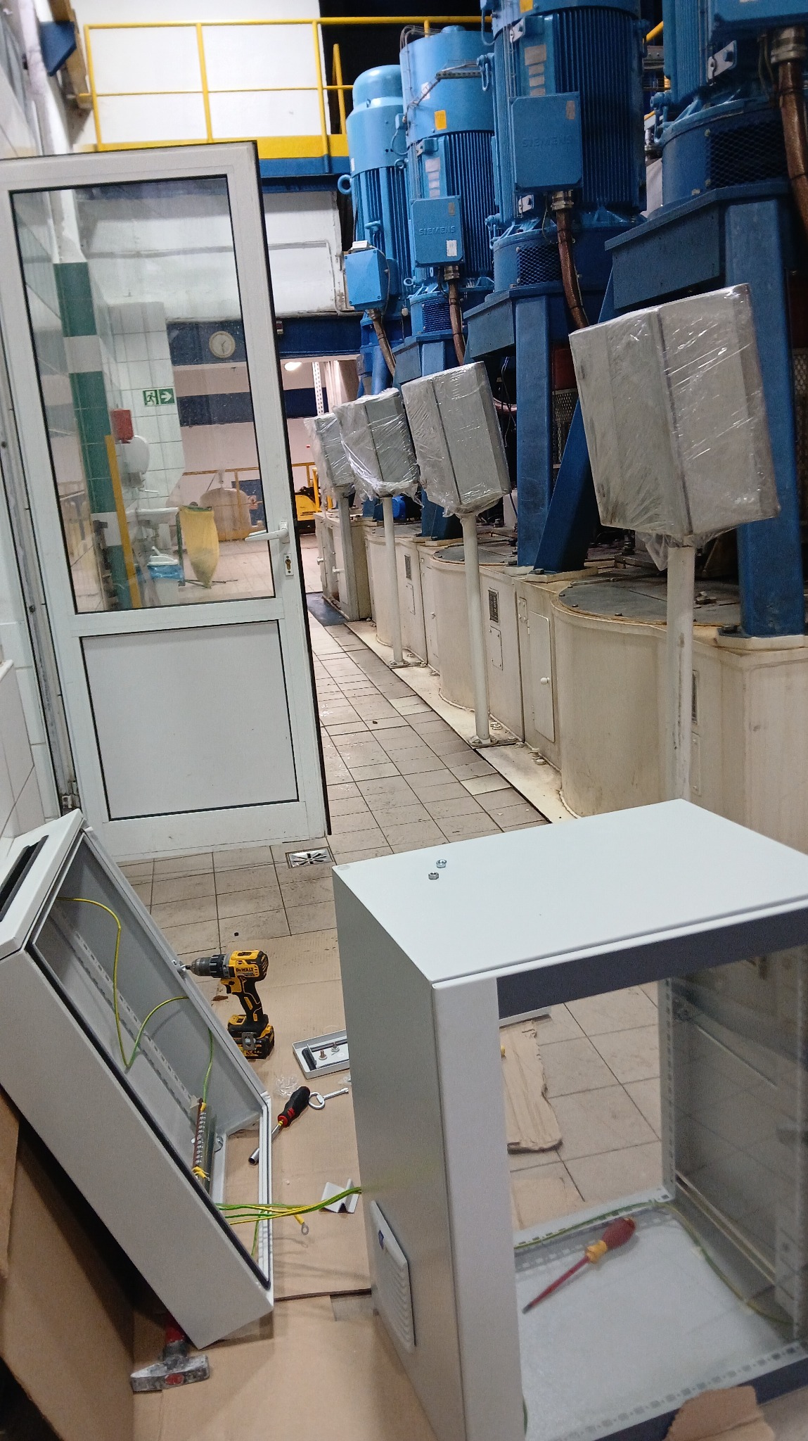 Rozdzielnice elektryczne w trakcie montażu w hali przemysłowej z rzędem silników. Widoczne narzędzia: wkrętarka, śrubokręty. Częściowo widoczna instalacja elektryczna.