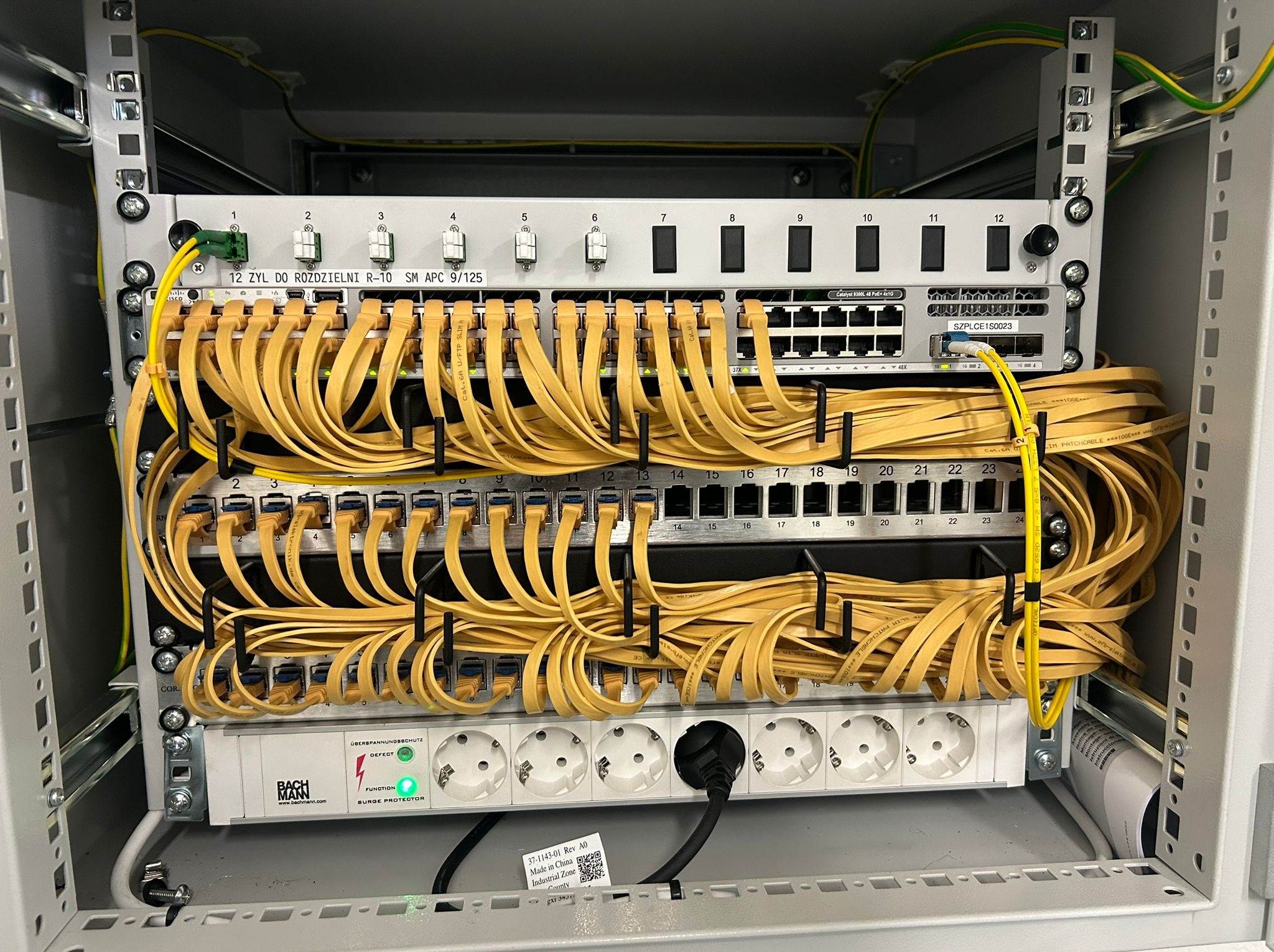 Szafa rack z patch panelem, switchami i listwą zasilającą Bachmann. Żółte kable patch cord uporządkowane w uchwytach. Widoczne porty RJ45 i złącza światłowodowe.